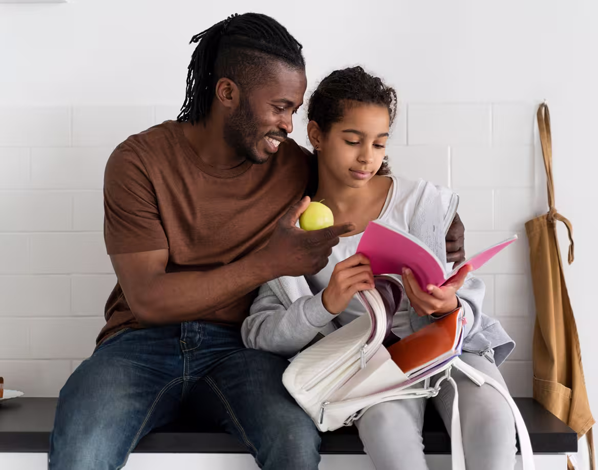 Man giving a green apple to a girl reading a pink notebook, sitting together indoors.