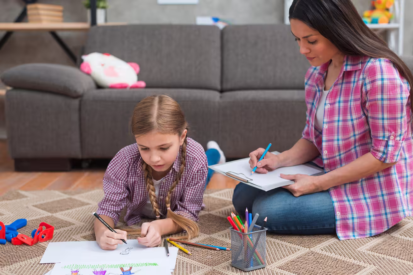 Woman and girl drawing with colored pencils on carpet in living room with gray sofa in background.