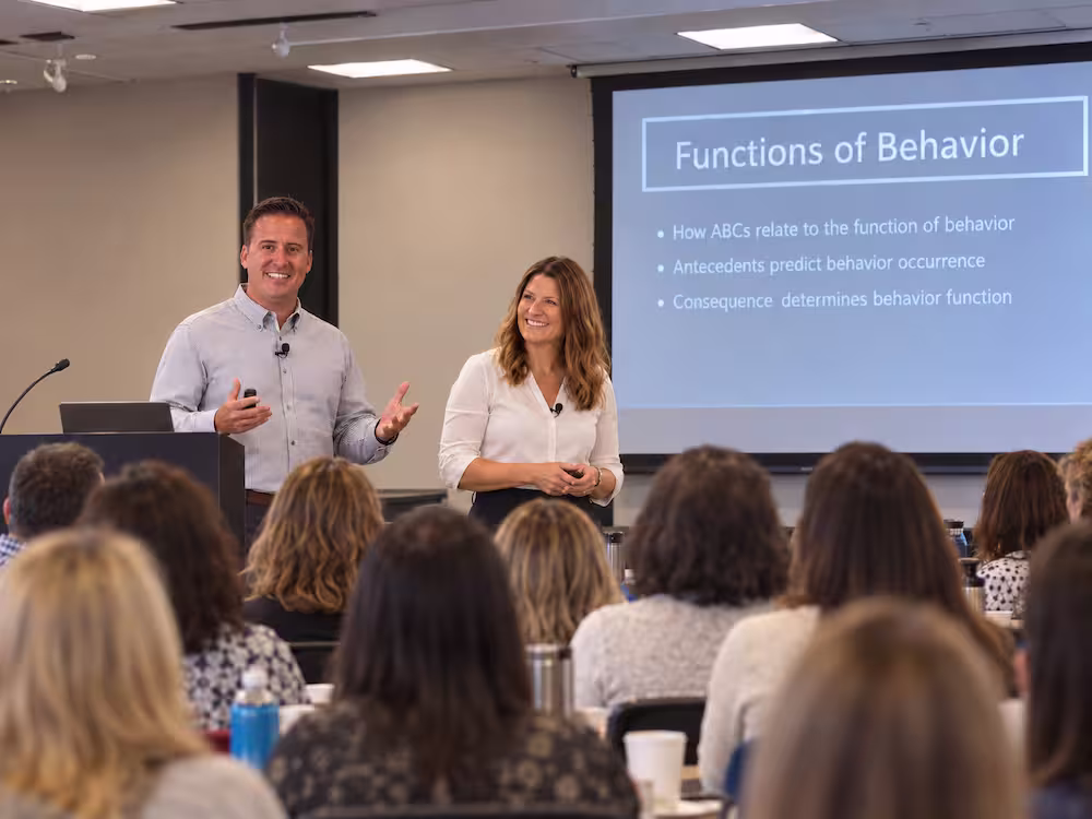 Man and woman standing at the front of a room giving a presentation to an audience, with a screen behind them showing a slide titled 'Functions of Behavior' listing bullet points about behavior functions.