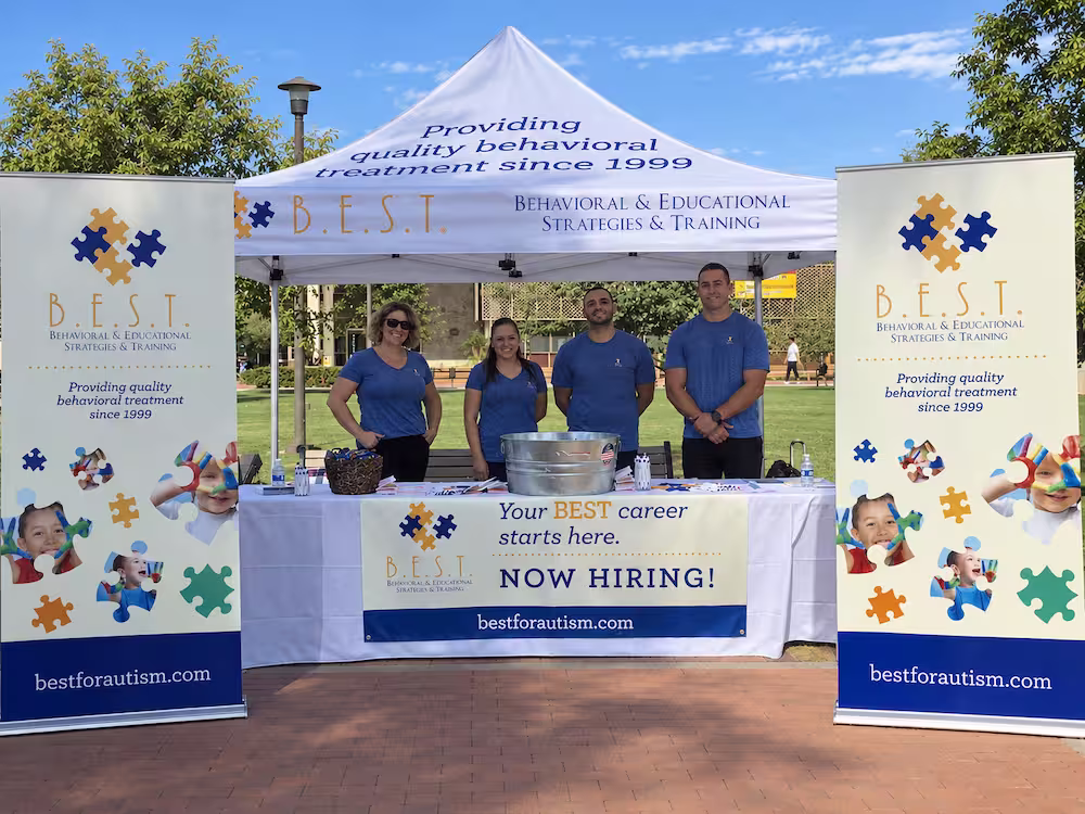 Four people wearing blue shirts stand behind a table under a white canopy tent with the text 'Providing quality behavioral treatment since 1999' and 'B.E.S.T. Behavioral & Educational Strategies & Training.' Two vertical banners with puzzle piece designs and images of children flank the setup, and a banner on the table reads 'Your BEST career starts here. NOW HIRING!' with the website bestforautism.com.