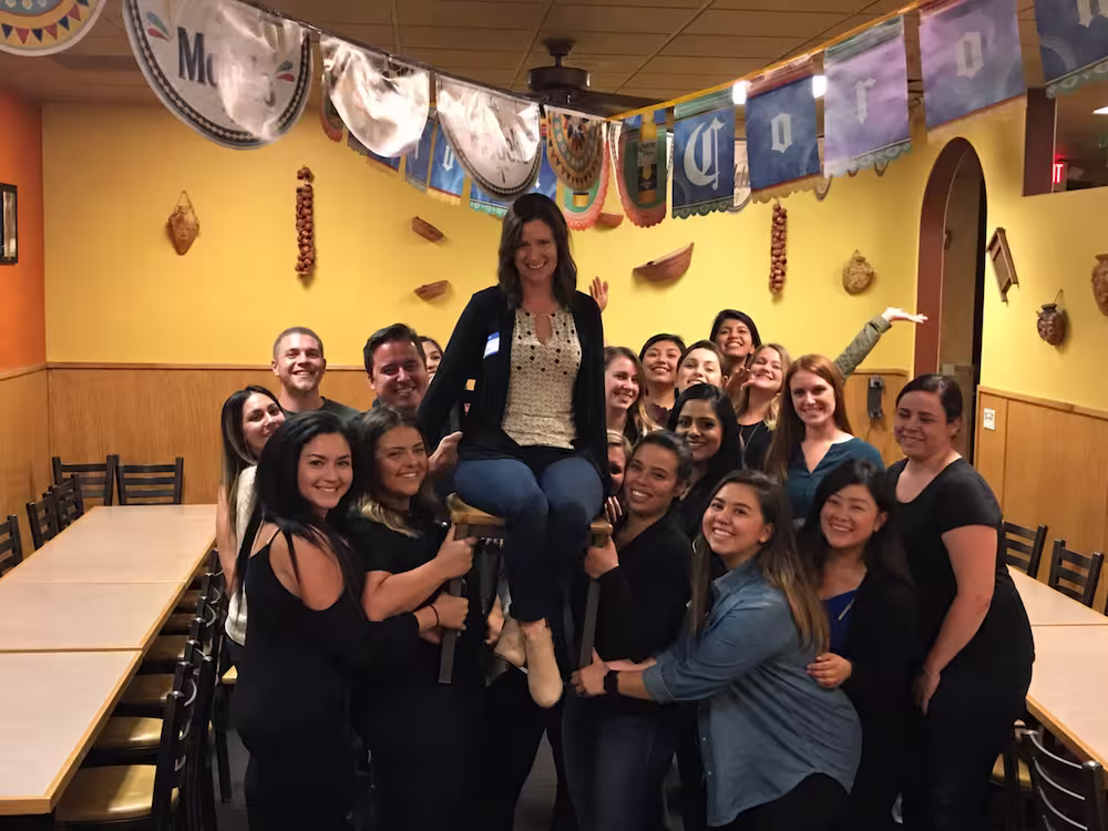 Group of smiling people indoors in a restaurant with yellow walls and hanging decorations, holding a woman seated on a chair lifted above the ground.