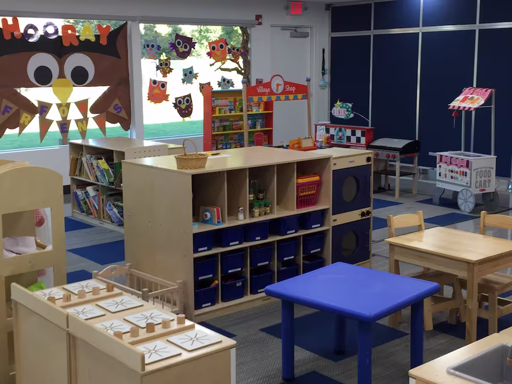 Bright, well-organized preschool classroom with small tables and chairs, colorful storage bins, educational toys, and owl-themed decorations on the windows, including a large brown owl with the word 'HOORAY' and smaller owl cutouts hanging above.