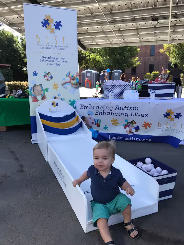 A young child sitting at the bottom of a white ball toss game set up outdoors with banners promoting B.E.S.T. Behavioral & Educational Strategies & Training, emphasizing support for autism and enhancing lives.