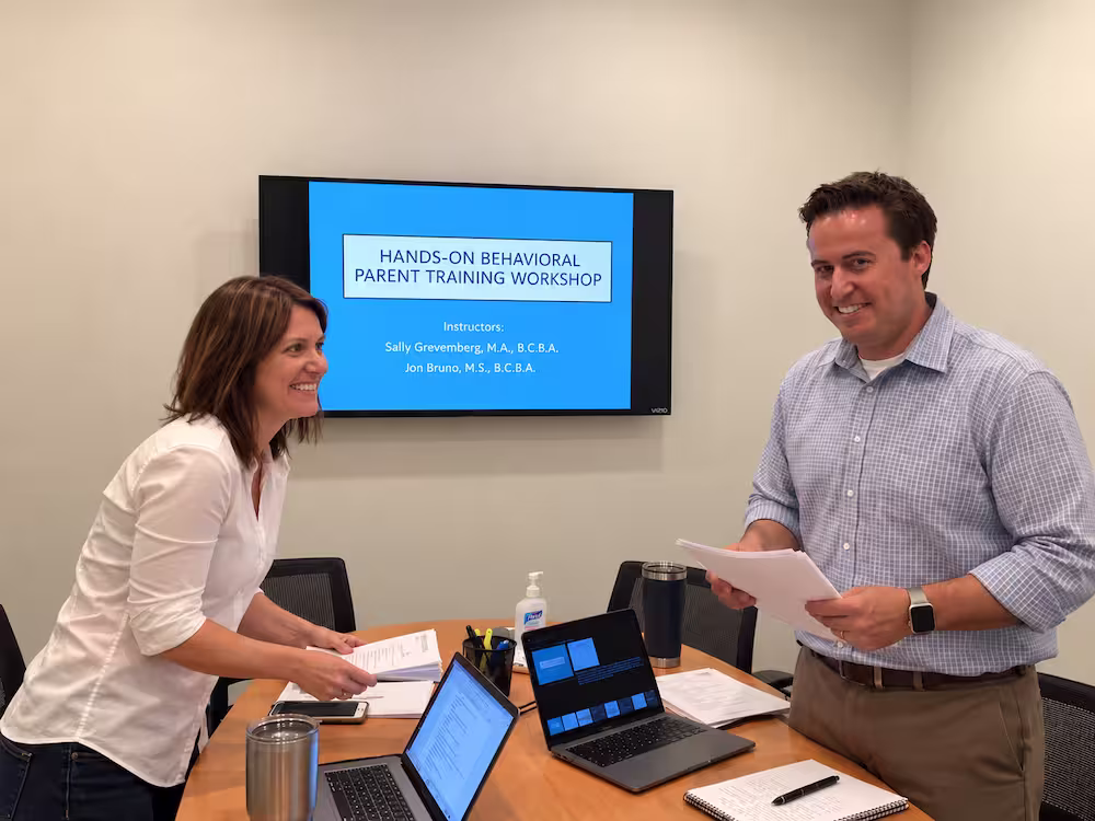 Two instructors smiling and standing in a meeting room with laptops, papers, and a sanitizer bottle on the table, beneath a screen displaying 'Hands-On Behavioral Parent Training Workshop' with instructor names.