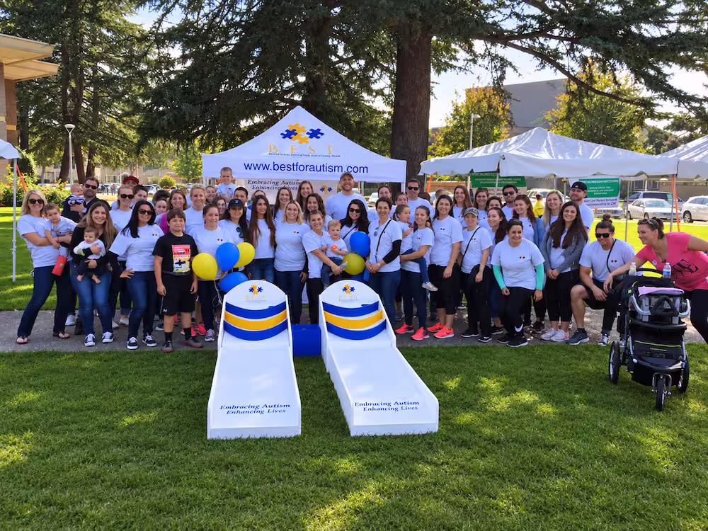 A large group of people standing outdoors on grass in front of a white tent with the website bestforautism.com, posing for a group photo during an autism awareness event. Two cornhole boards with blue and yellow stripes and the phrase 'Embracing Autism Enhancing Lives' are set up in front of the group, with some participants holding blue and yellow balloons.