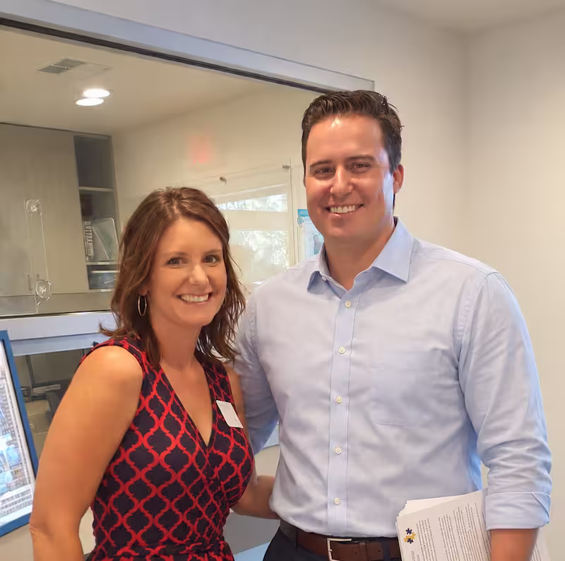 Smiling woman in a patterned sleeveless dress stands with smiling man in light blue button-up shirt holding folded papers in a modern indoor setting.