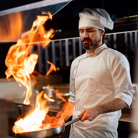 Chef in white uniform and hat cooking with flames in a professional kitchen.