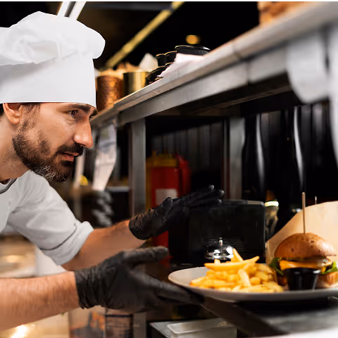 Chef in white uniform and black gloves placing a plate with a burger and French fries on a pass-through counter in a kitchen.