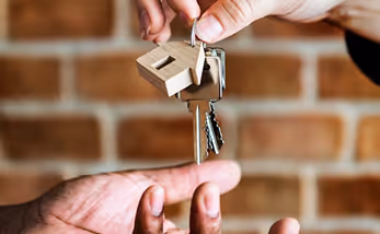 Hand holding a keychain with a small wooden house and keys, handing it to another outstretched hand.