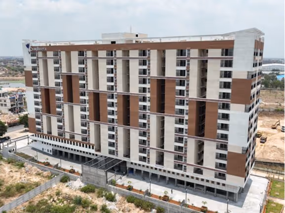 A multi-story residential building under construction with brown and beige vertical panel design, surrounded by fencing and some vegetation.