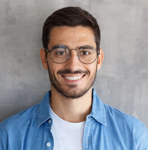 Smiling young man with round glasses wearing a blue shirt against a gray textured wall.
