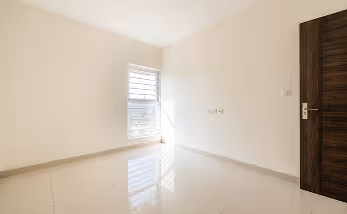 Empty room with beige tiled floor, white walls, a wooden door, and sunlight streaming through a small window.