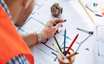 Person wearing an orange vest drawing on architectural blueprints with colored pencils and a ruler nearby.