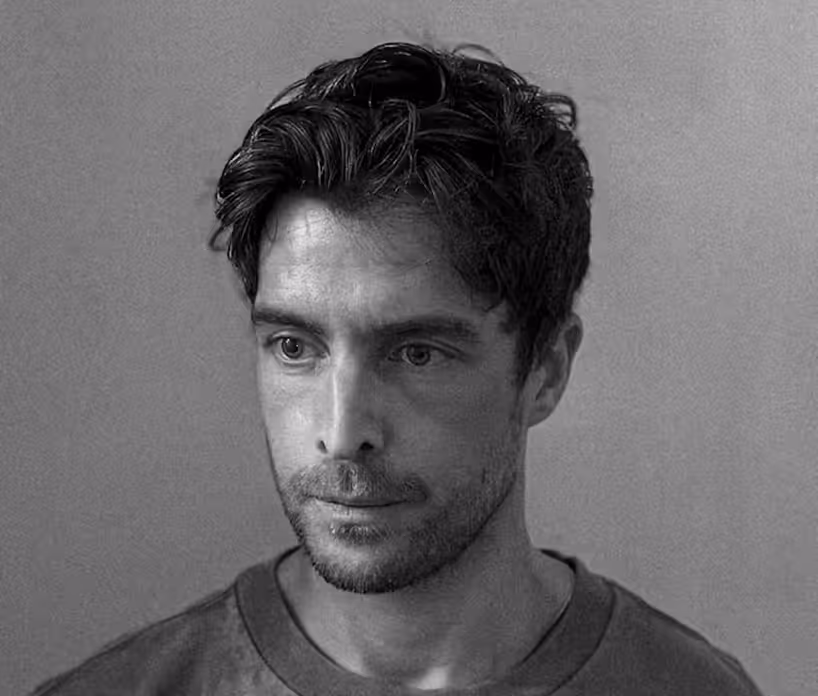 Black and white close-up of a man with short dark hair and stubble, wearing a plain shirt.