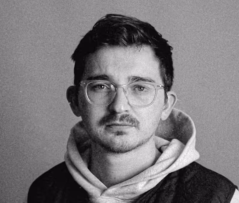 Man with glasses and facial hair wearing a hoodie and jacket looking directly at the camera.