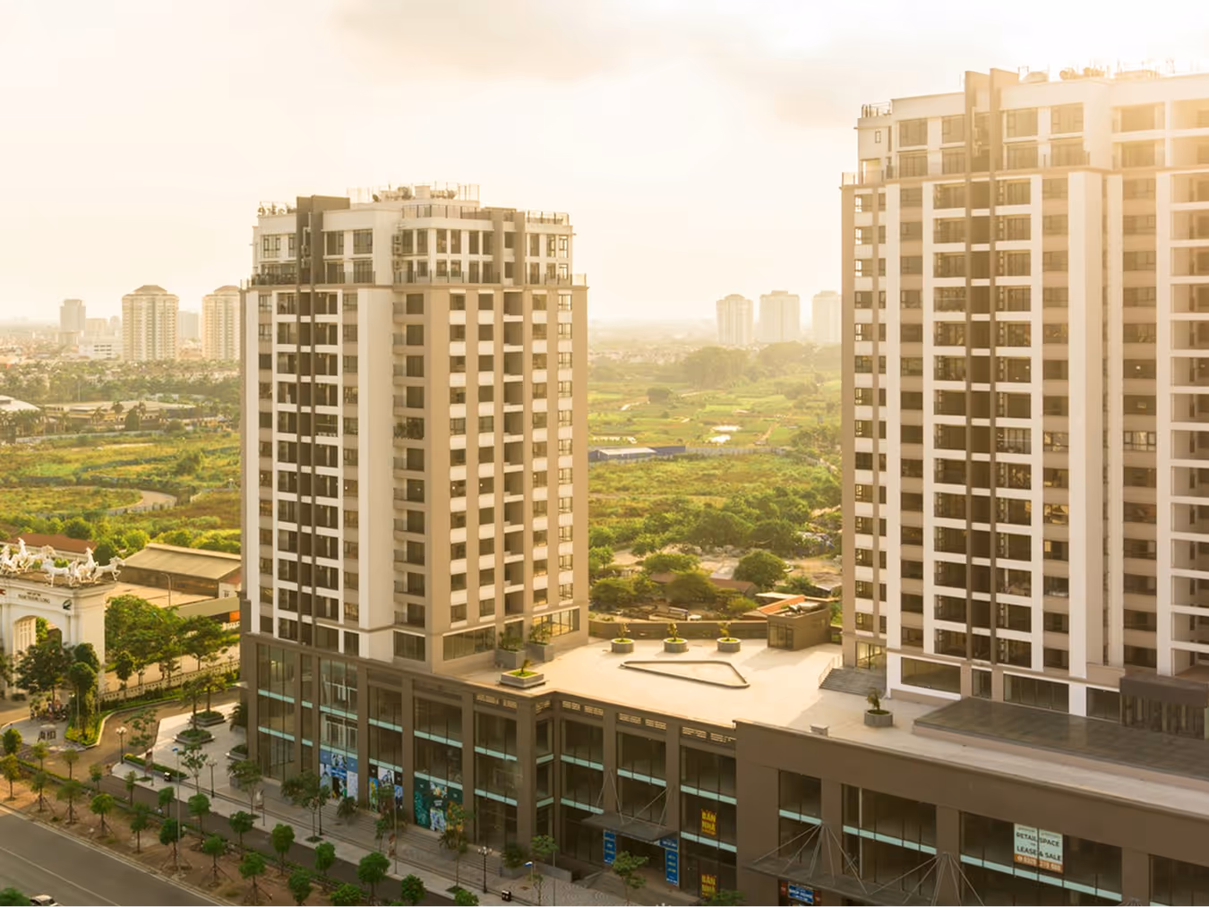 Two modern high-rise buildings with glass and concrete facades amid green fields at sunset.