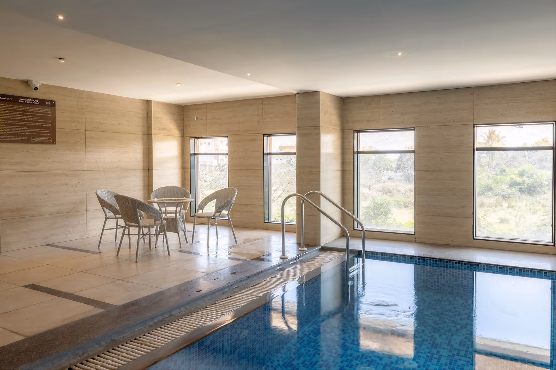 Indoor swimming pool with blue tiles, metal railing, and wicker chairs with a small table by windows.