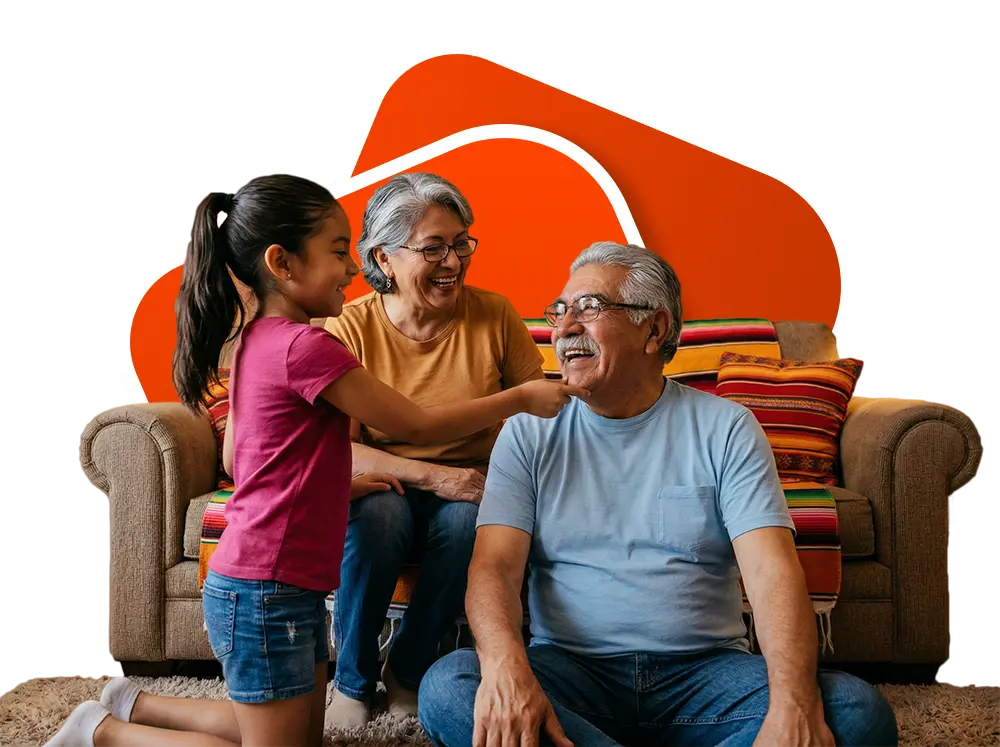 Smiling grandparents seated on a couch with colorful blanket and pillows, interacting joyfully with their granddaughter who is kneeling in front of them.