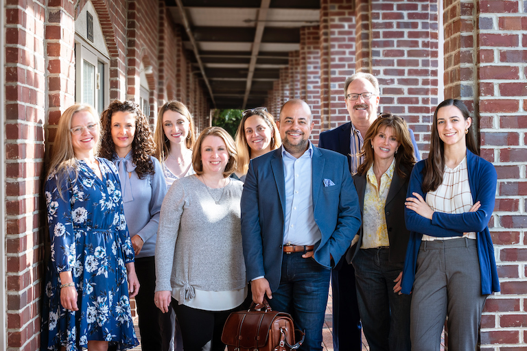 The nine people team at Victor Medina's company pose side-by-side in an outdoor scene. They are wearing shades of blue and gray and smiling.