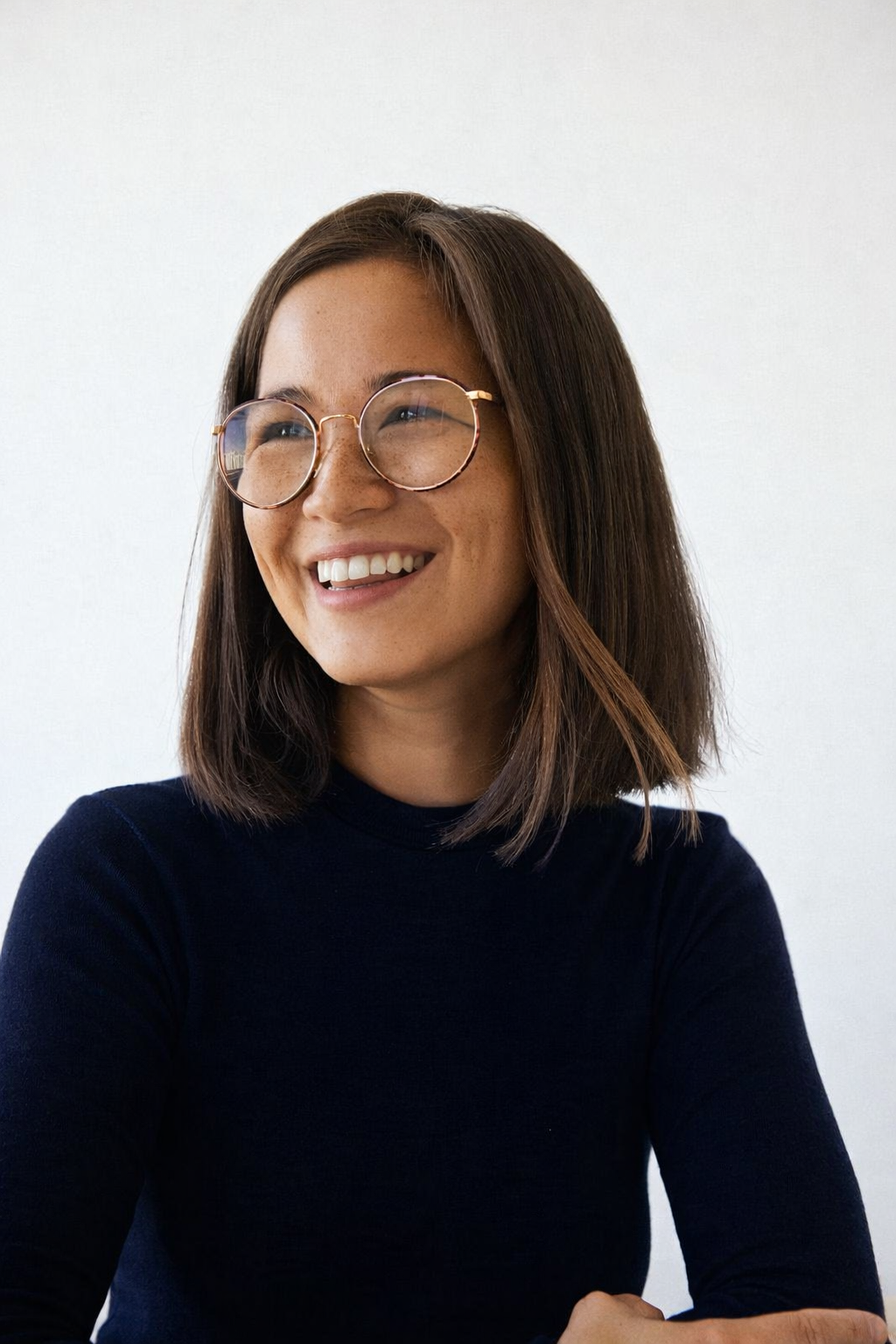 Young woman with shoulder-length brown hair and round glasses smiling against a plain background.