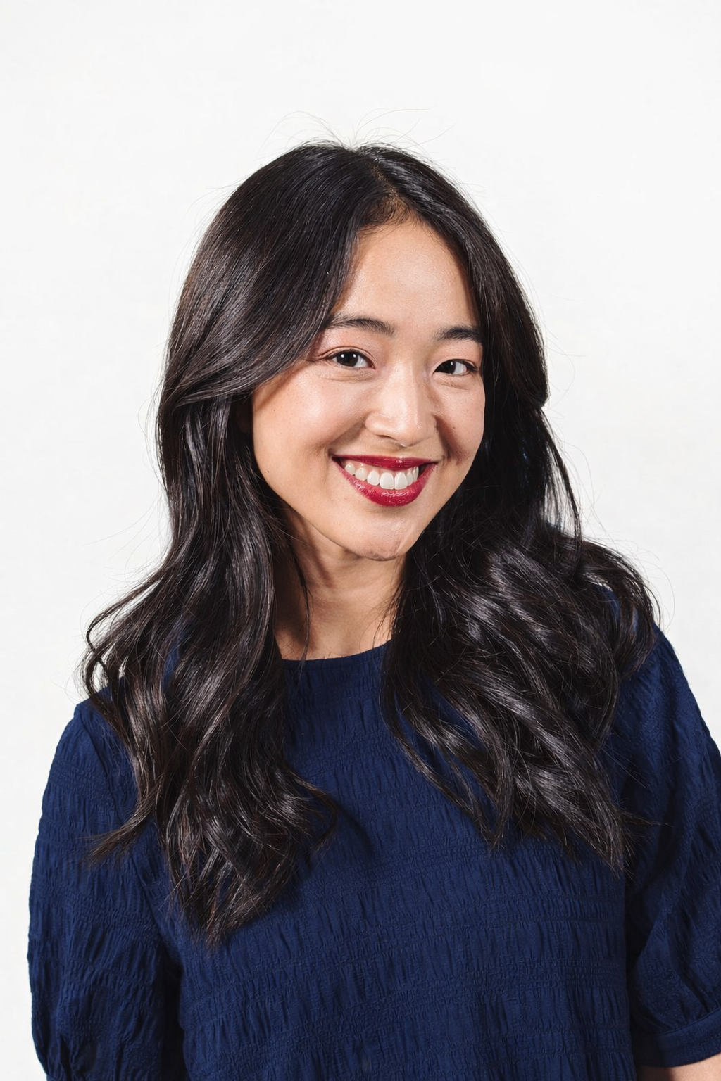 Smiling woman with long dark wavy hair wearing a navy blue textured top against a white background.