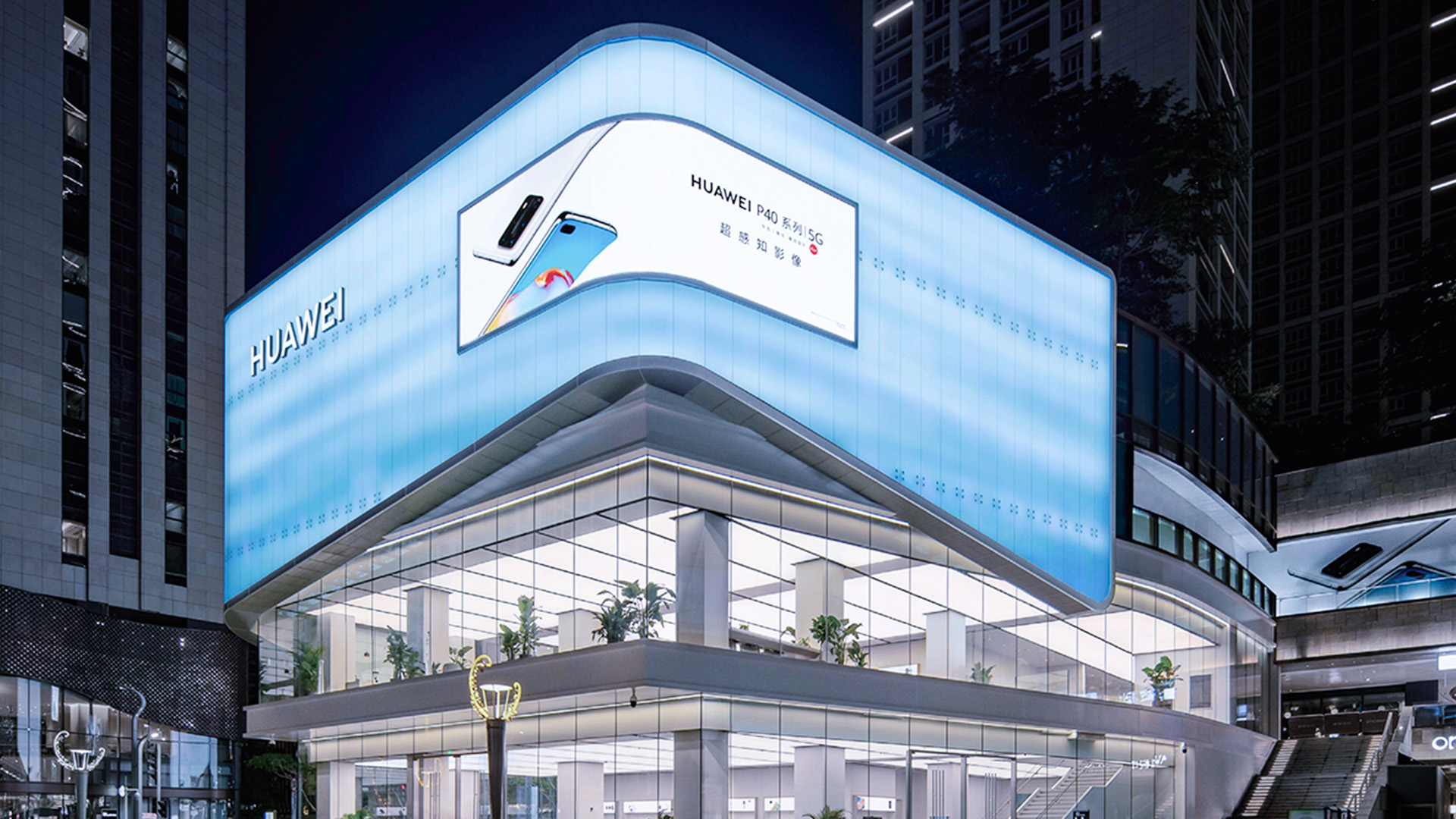 Modern Huawei store at night with large illuminated signage and glass walls showing interior plants.