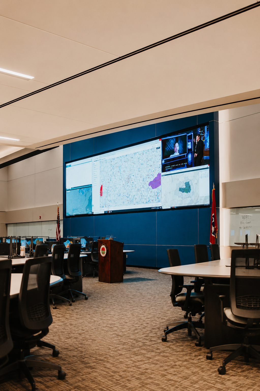 Empty control room with rows of chairs and a large multi-screen display showing maps and news.