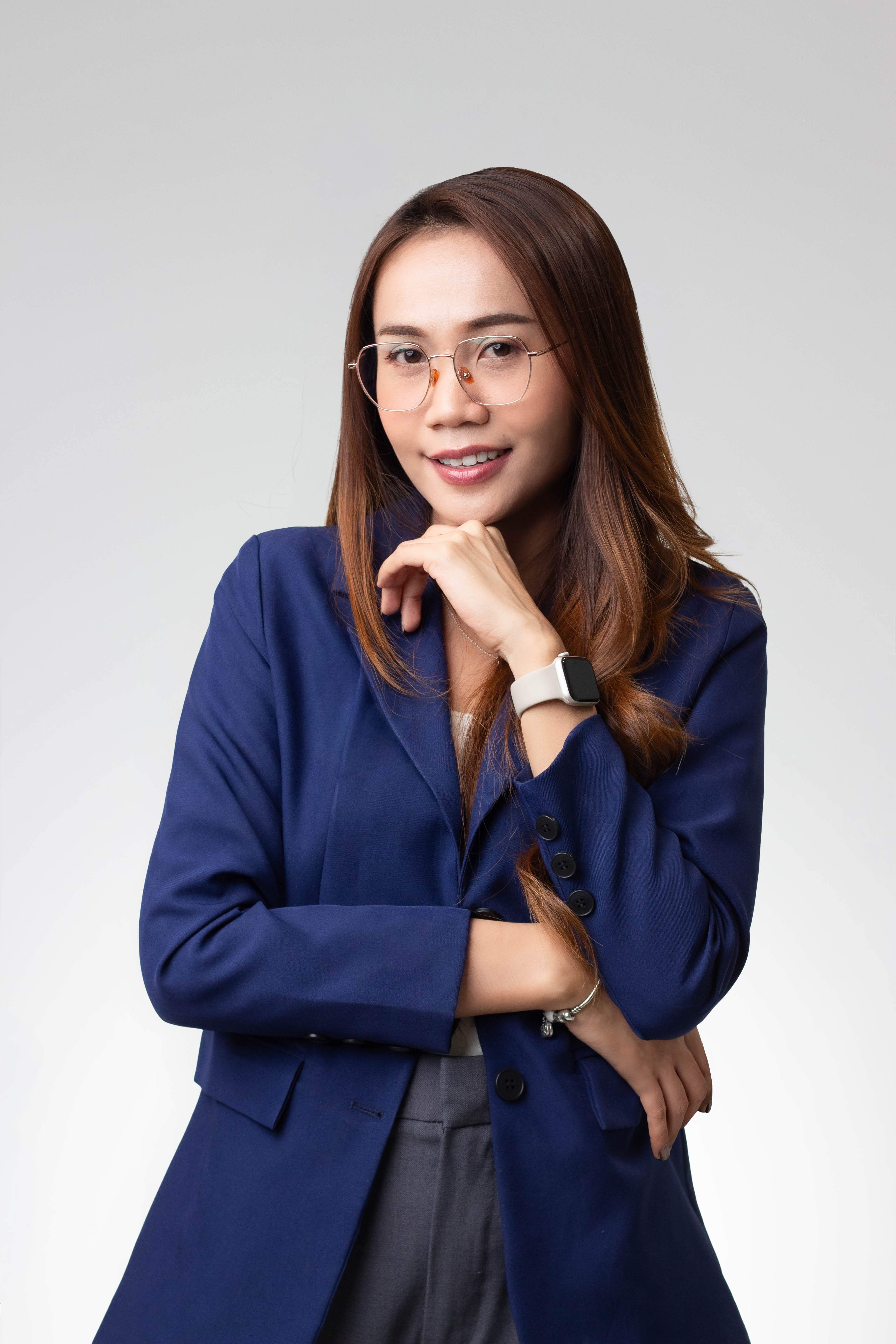 Smiling woman with long hair, glasses, blue blazer, and smartwatch posing against gray background.