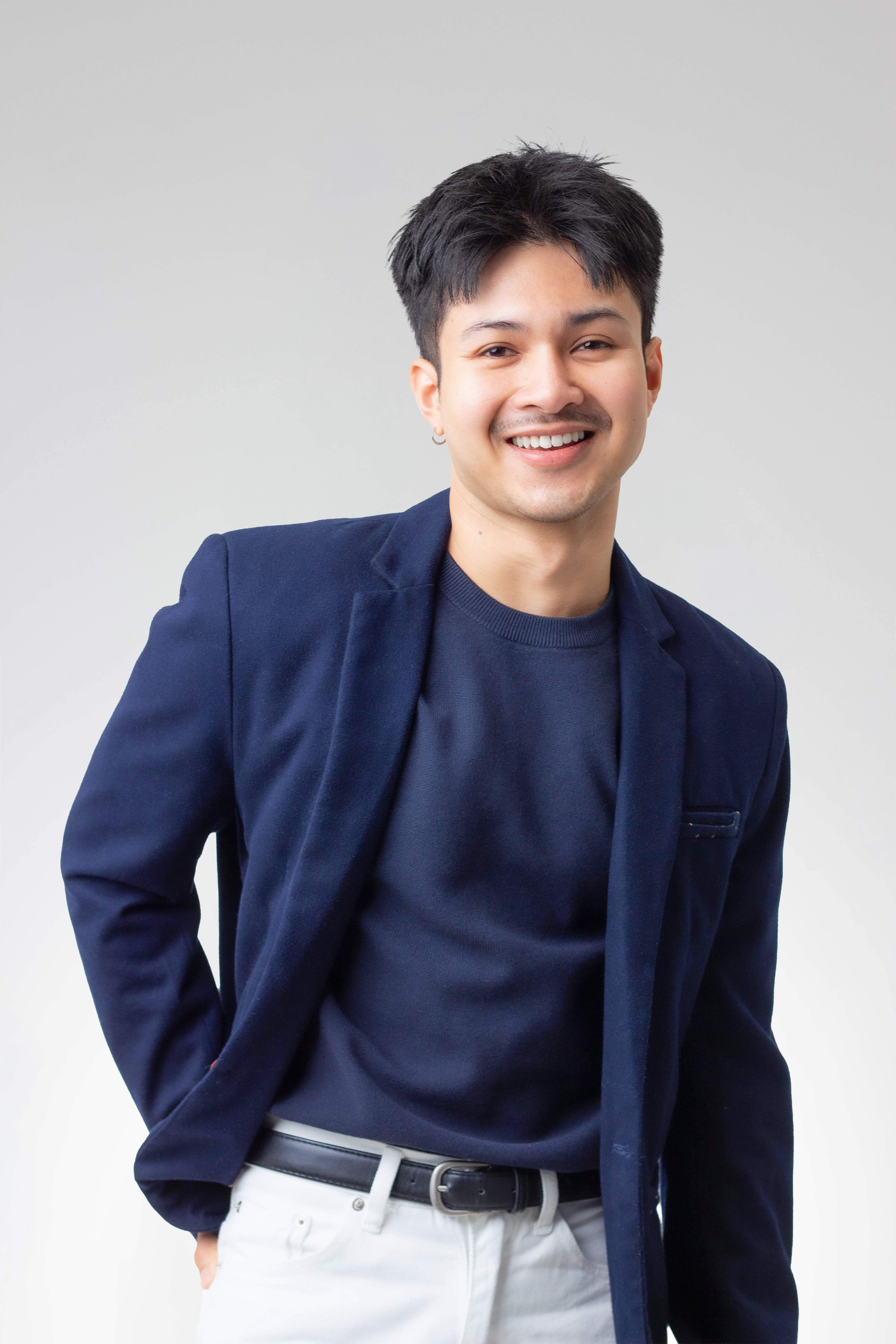 Smiling man in navy blazer and sweater with white pants and black belt against a plain background.