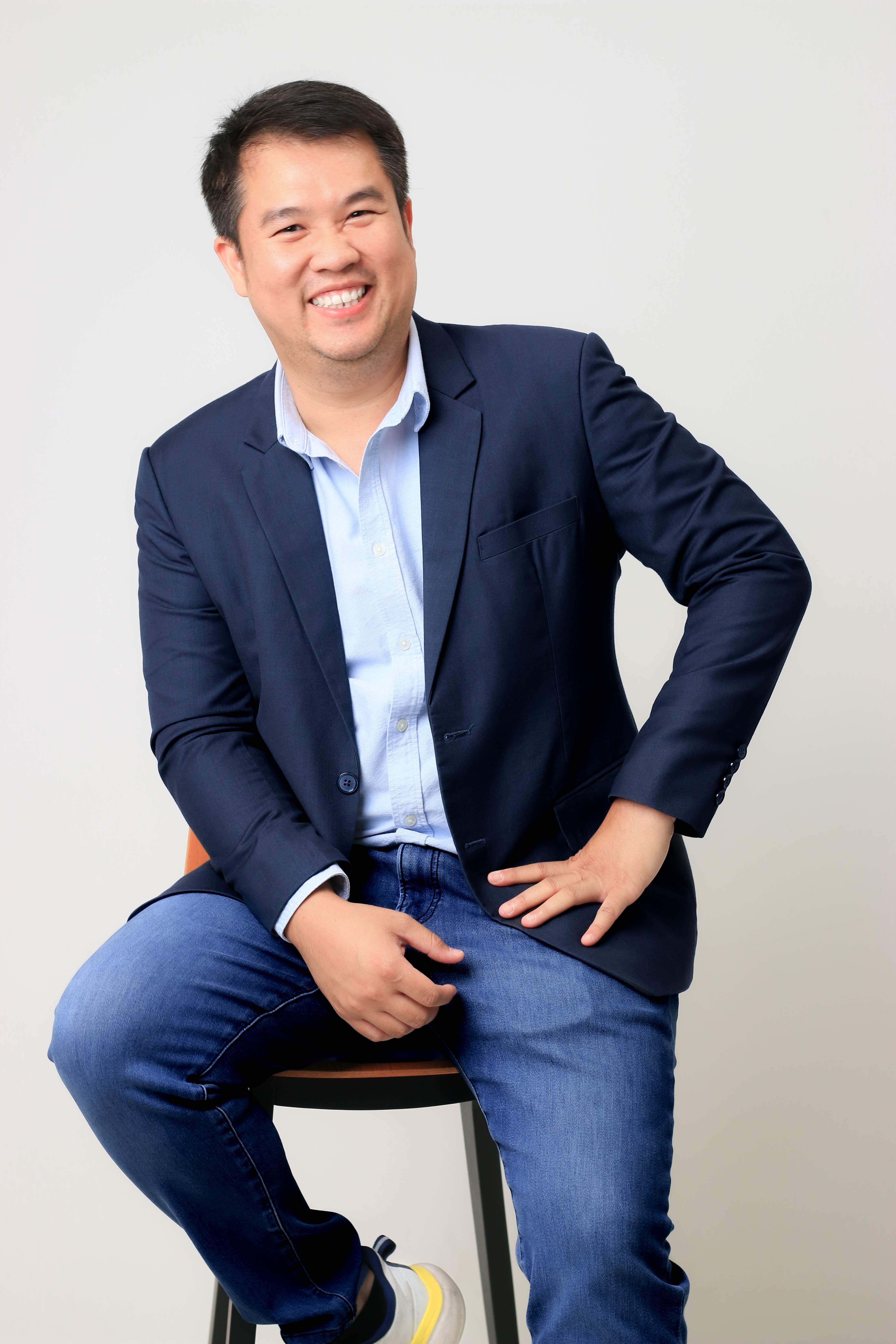 Smiling man sitting on stool wearing dark blazer, light blue shirt, jeans, and sneakers.