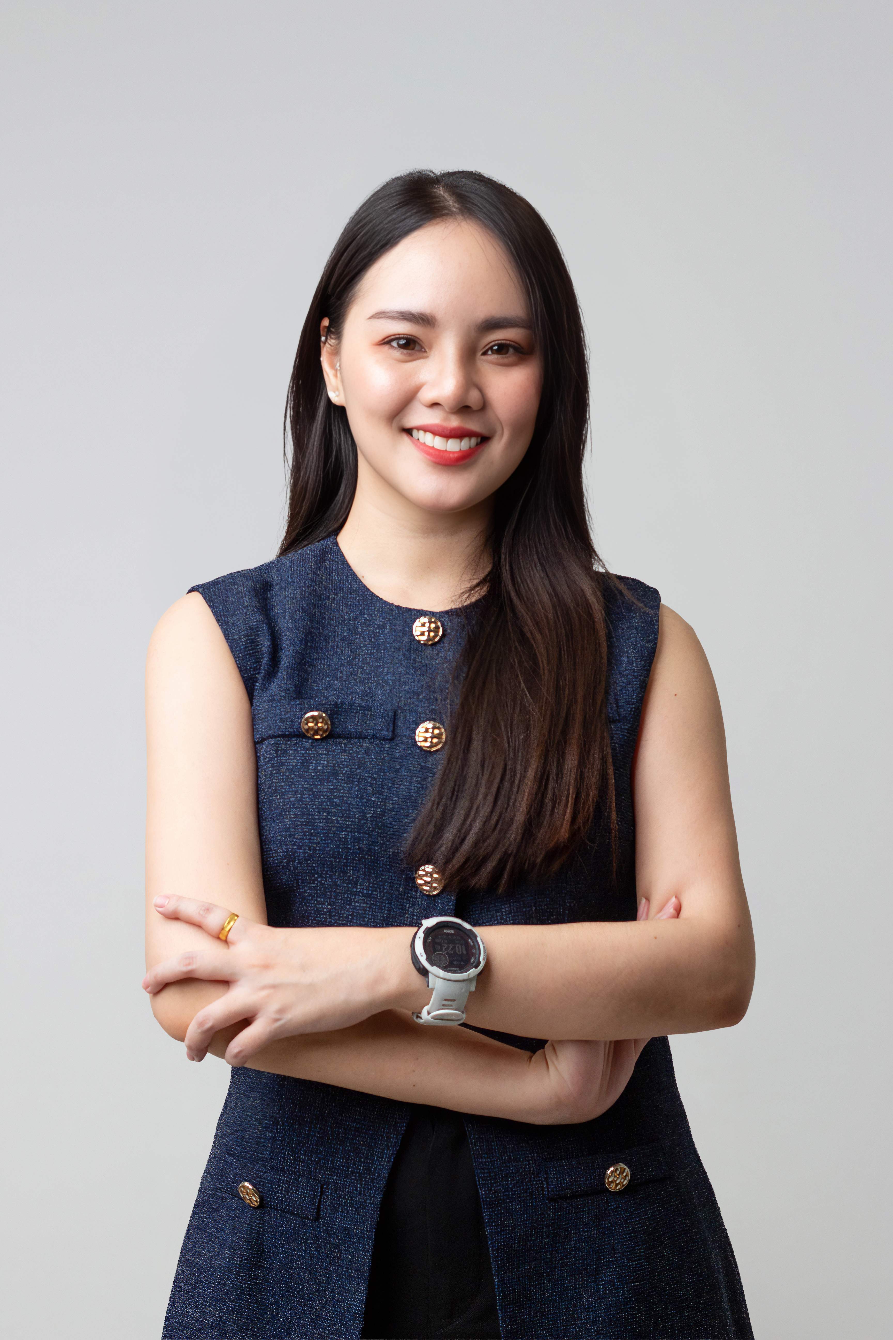 Smiling woman with long dark hair wearing a sleeveless blue vest with gold buttons and a white watch.