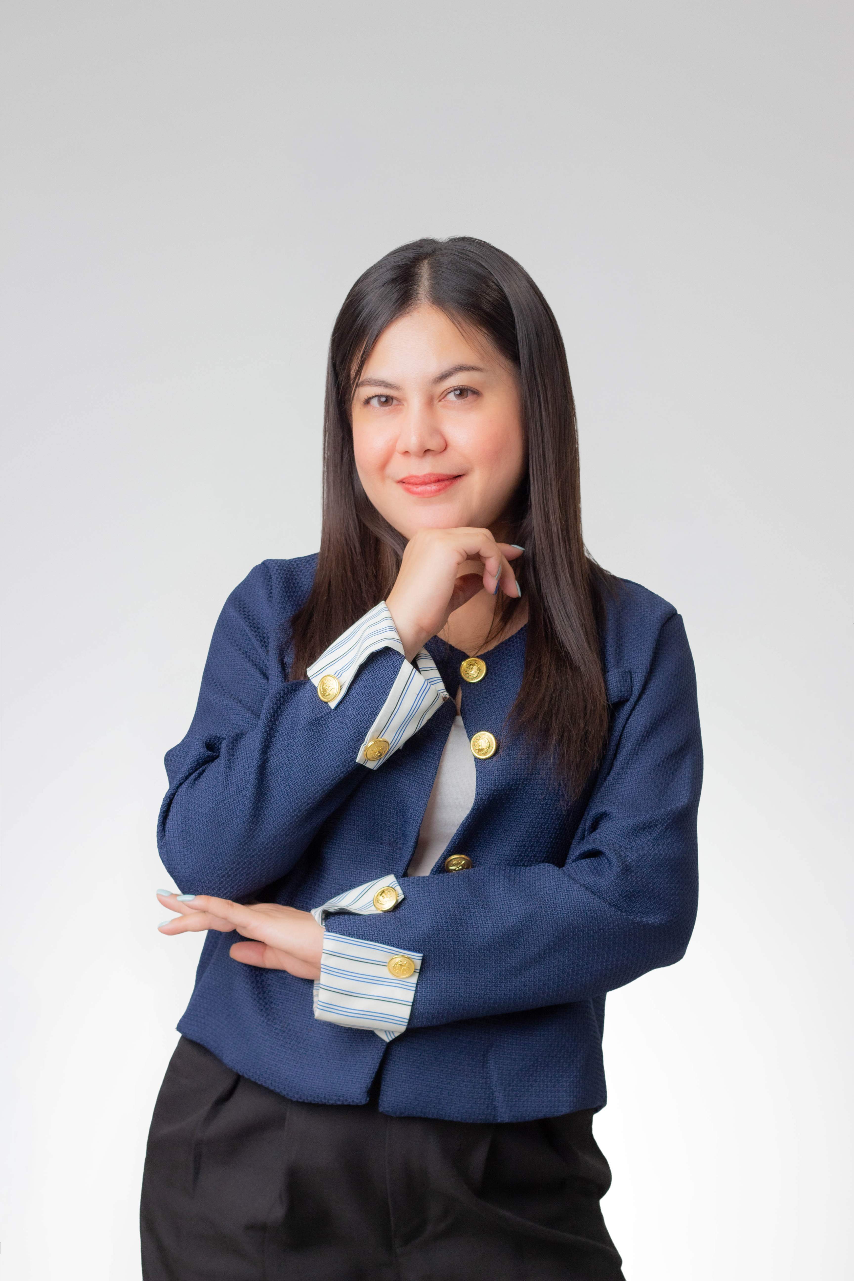 Woman with long dark hair in navy blazer with gold buttons posing confidently against a plain background.
