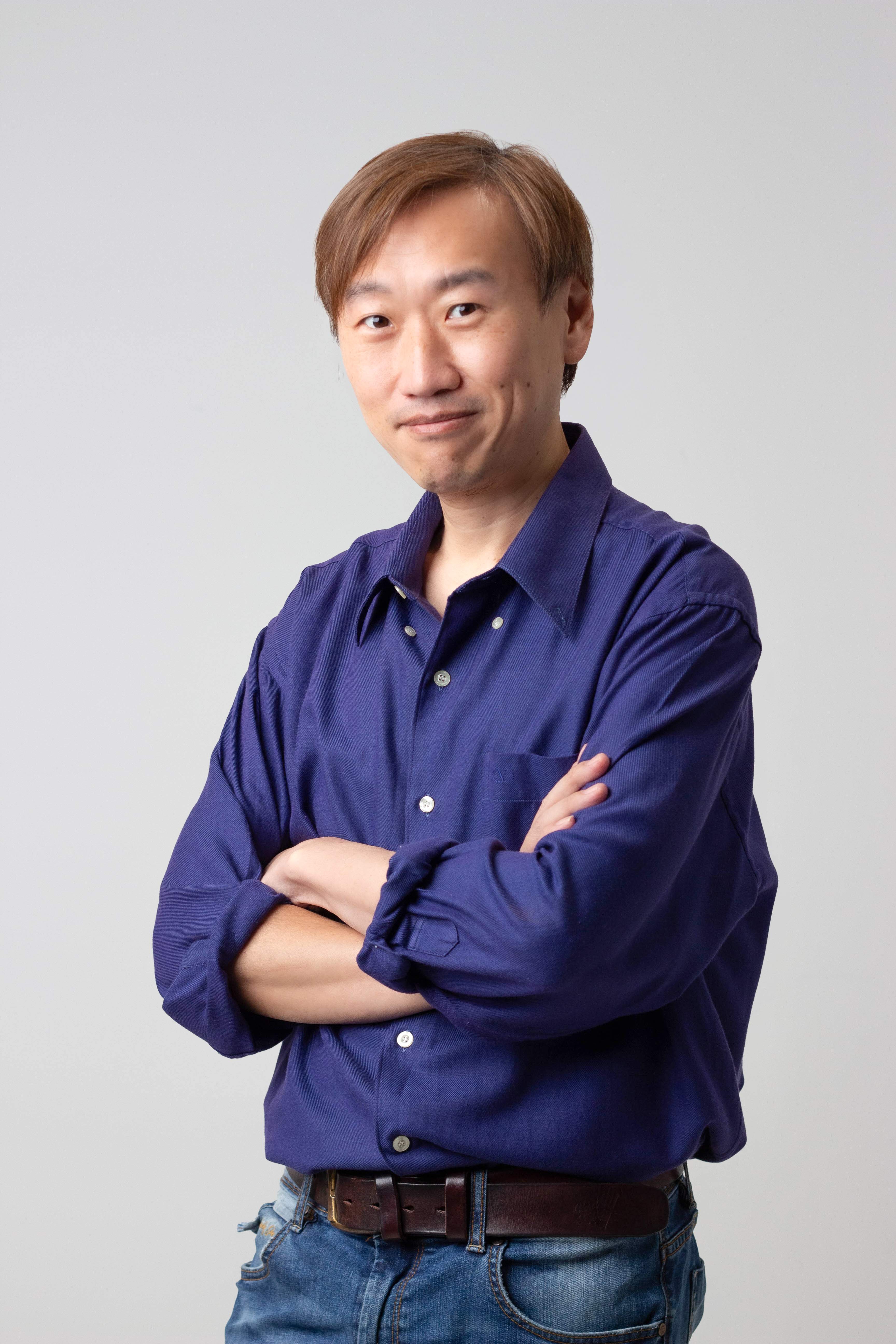 Man with light brown hair wearing a blue shirt and jeans with arms crossed, smiling slightly.