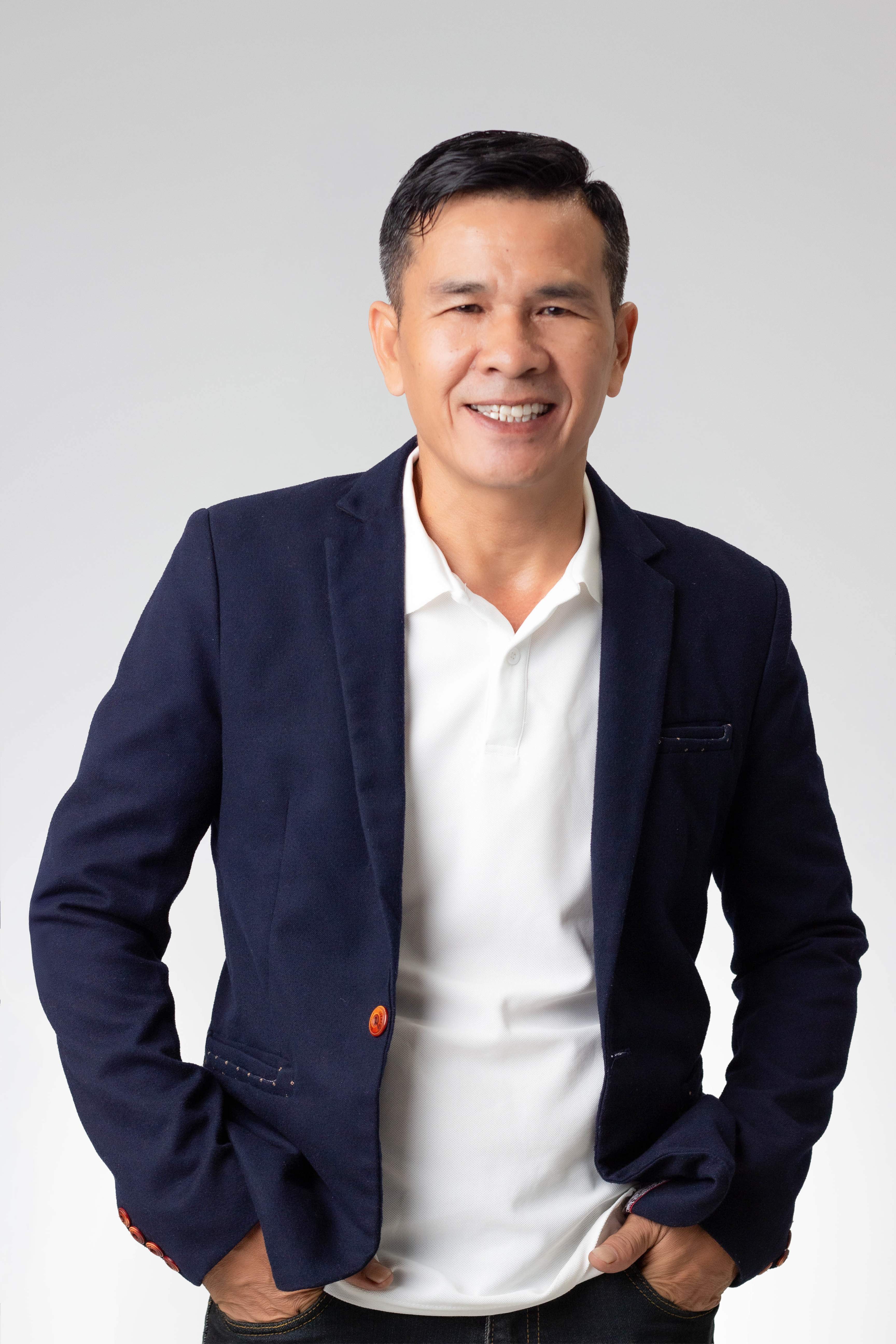 Smiling man wearing a navy blazer over a white polo shirt with hands in pockets, against a gray background.