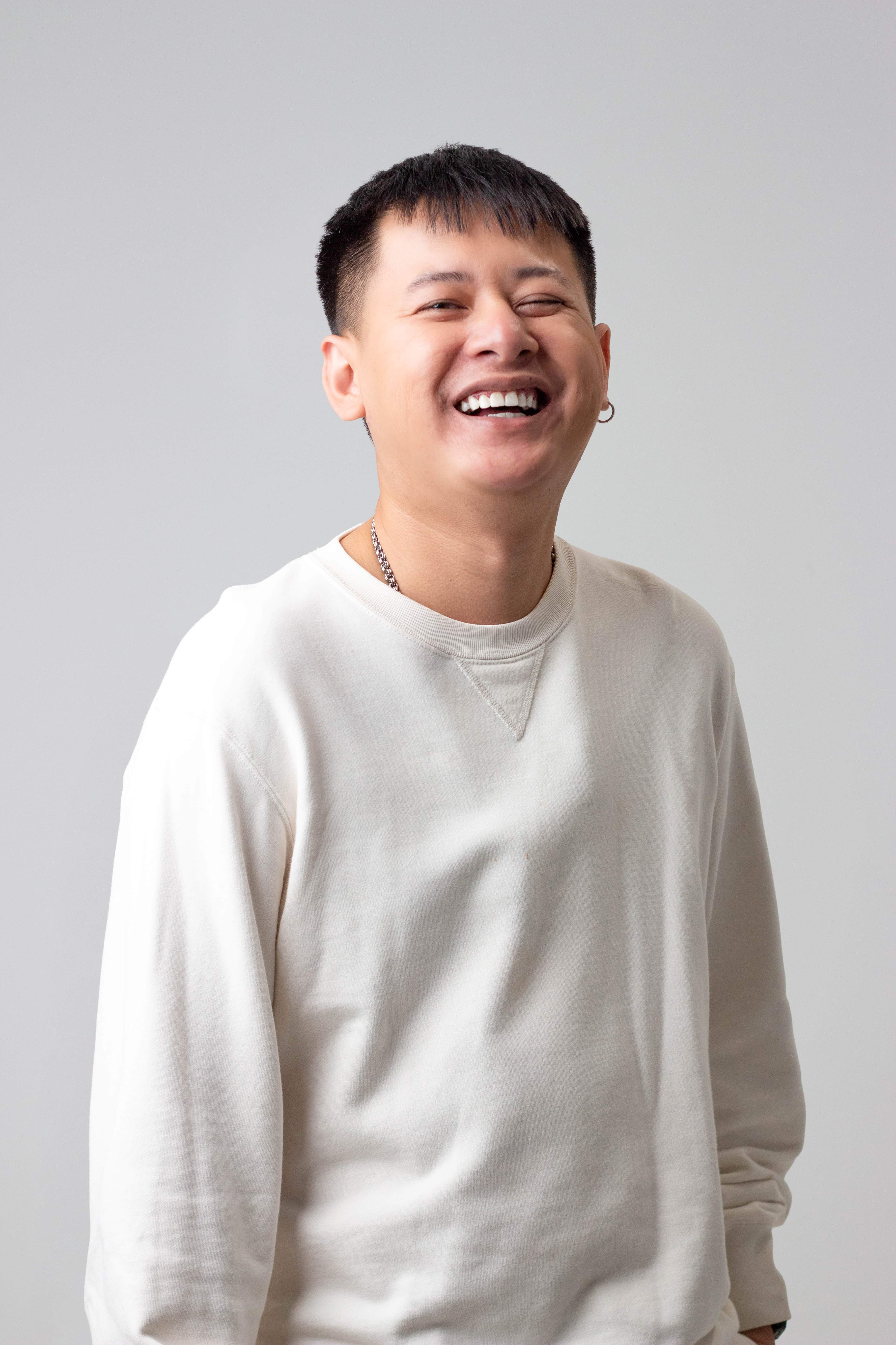 Smiling young man with short hair in white sweatshirt laughing against plain light background.