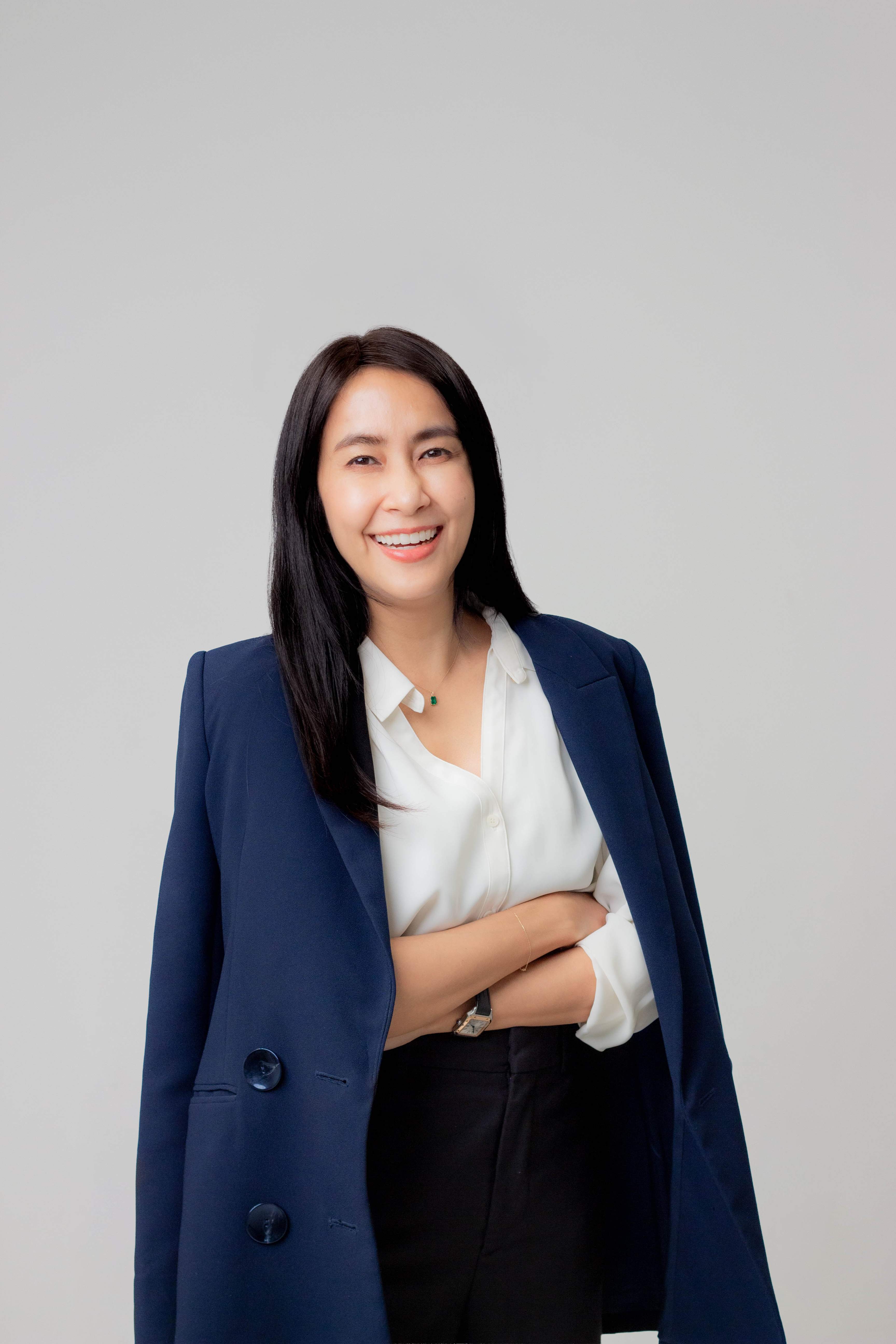 Smiling woman with long black hair wearing white blouse and navy blazer against gray background.