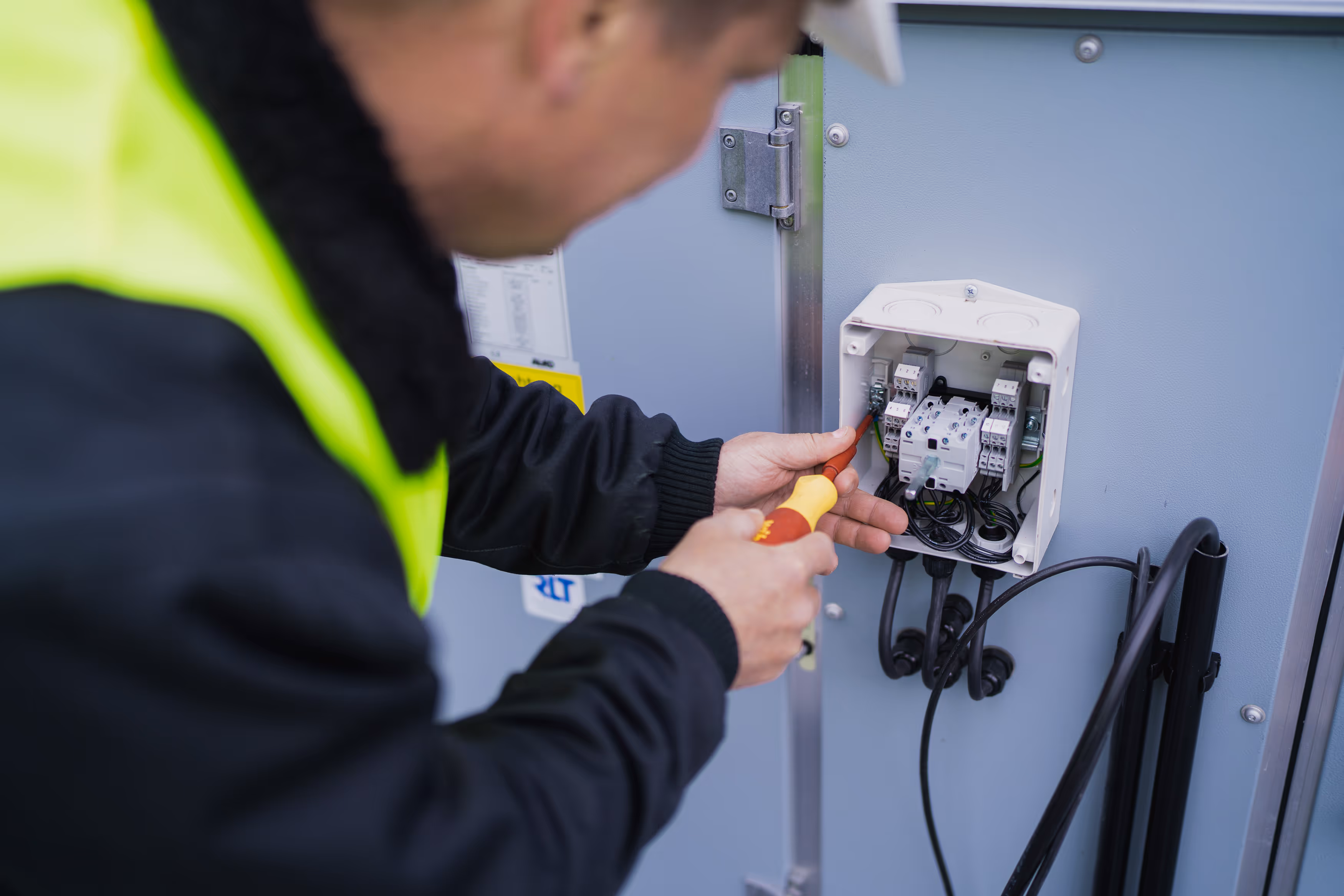 Elektriker mit gelber Warnweste und Schraubendreher arbeitet an offenem elektrischen Anschlusskasten.