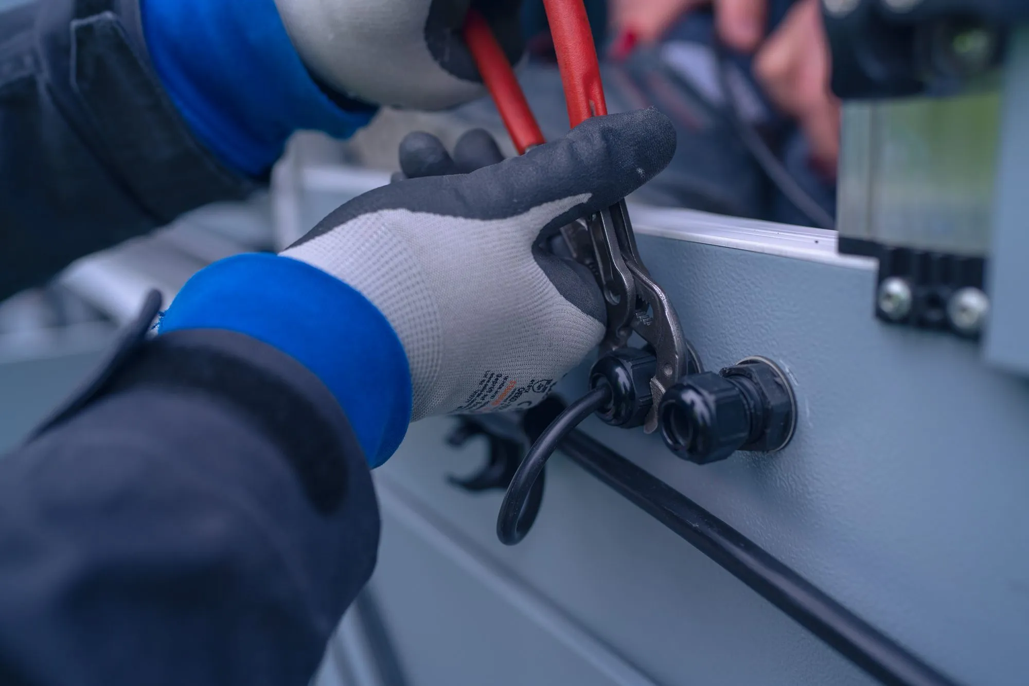 Hand in Arbeitshandschuh hält Zange und befestigt Kabel an einem grauen Metallgehäuse.