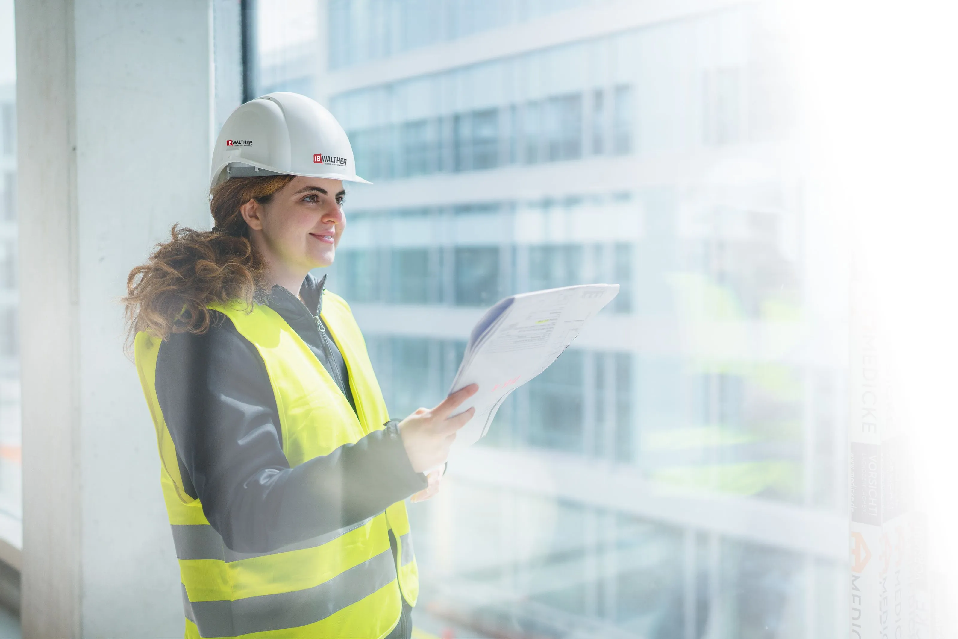 Mitarbeiterin von IB Walther mit gelben Sicherheitsweste und weißem Schutzhelm hält Baupläne und blickt lächelnd aus dem Fenster einer Baustelle.
