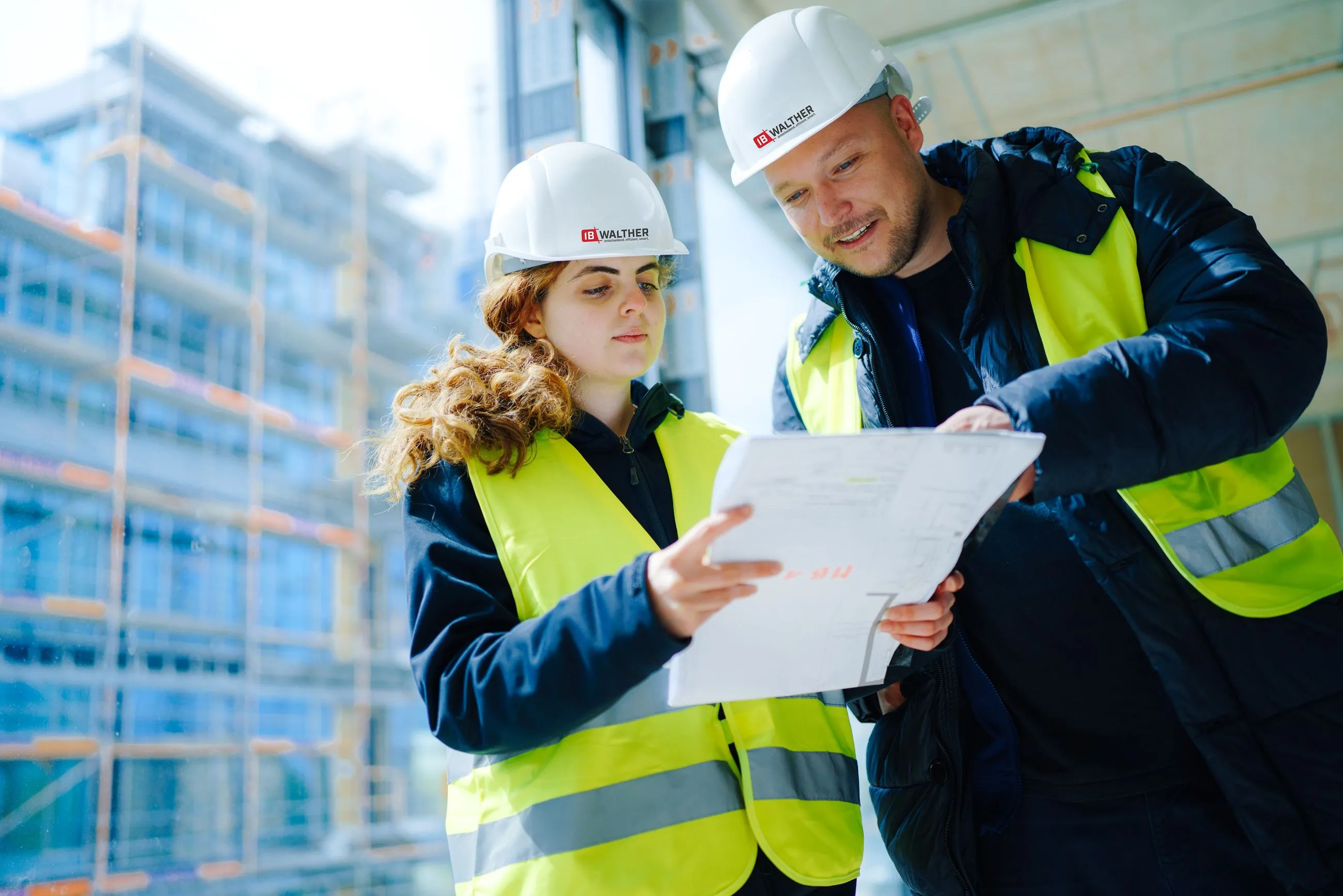Mitarbeiter:in von IB Walther mit weißen Helmen und gelben Warnwesten überprüfen einen Bauplan auf einer Baustelle.