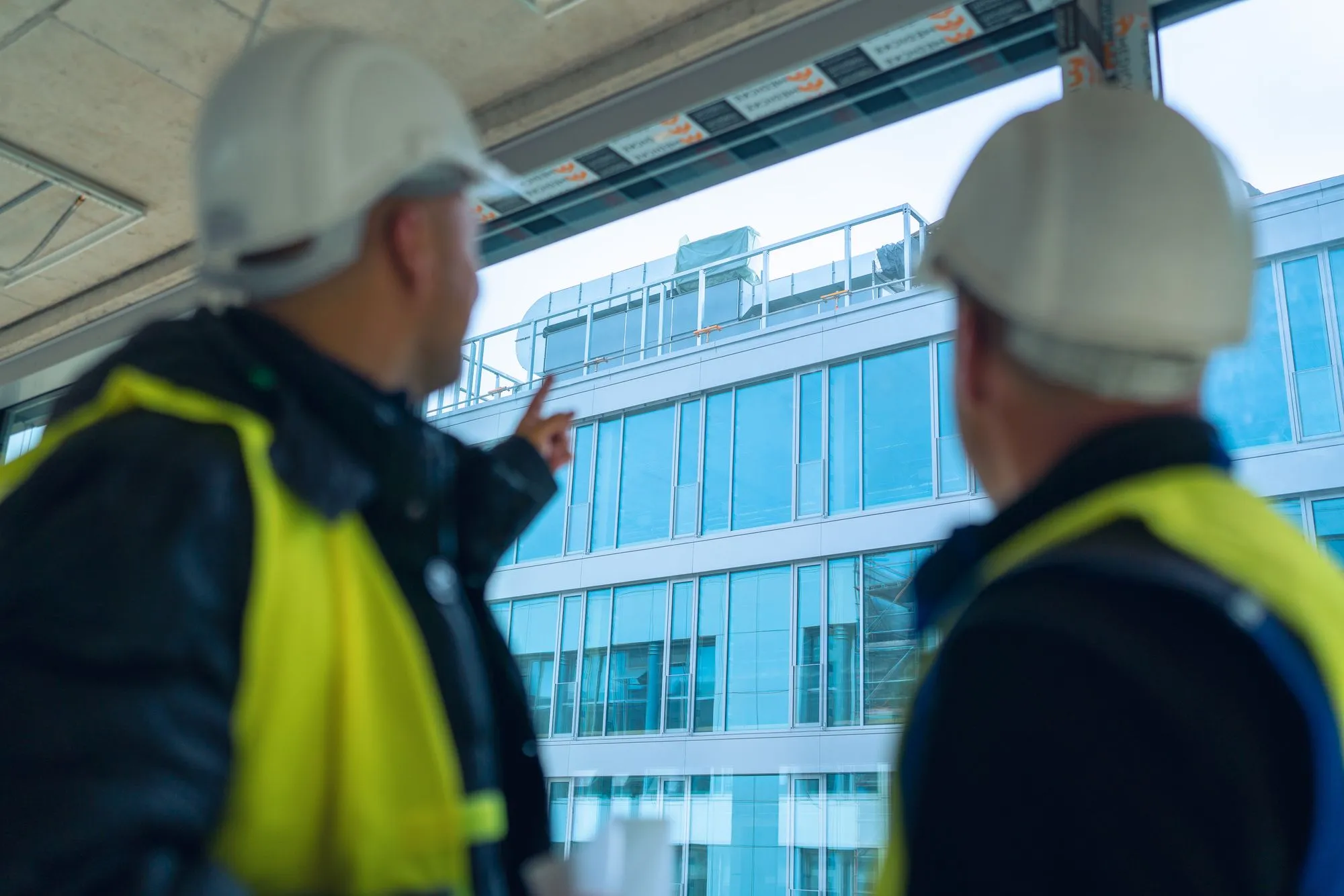 Zwei IB Walther Mitarbeiter mit weißen Helmen und gelben Sicherheitswesten betrachten und zeigen auf ein modernes Bürogebäude mit blauen Fensterfronten.
