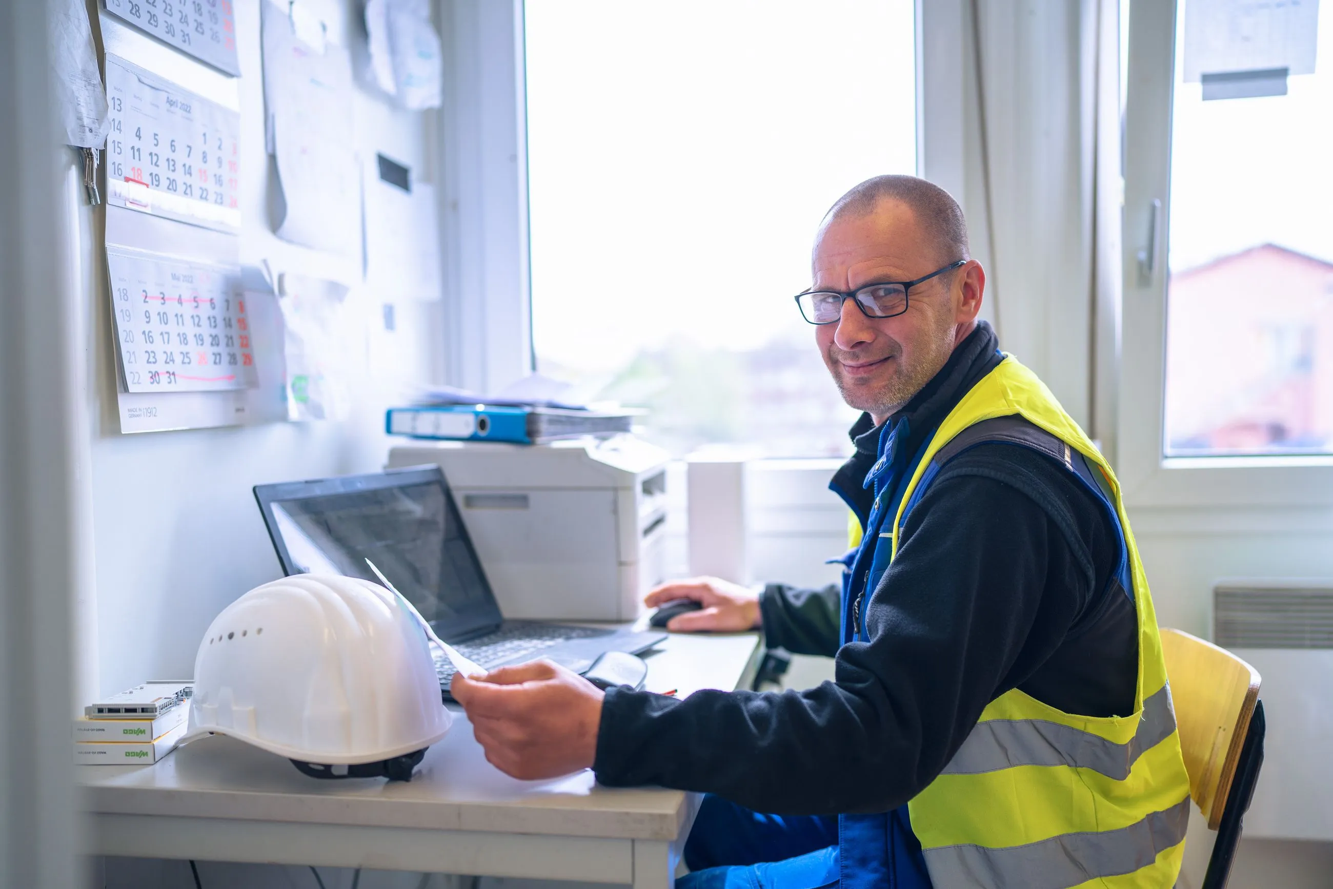 IB Walther Projektleiter mit reflektierender Weste arbeitet an einem Schreibtisch mit Laptop und Bauhelm.