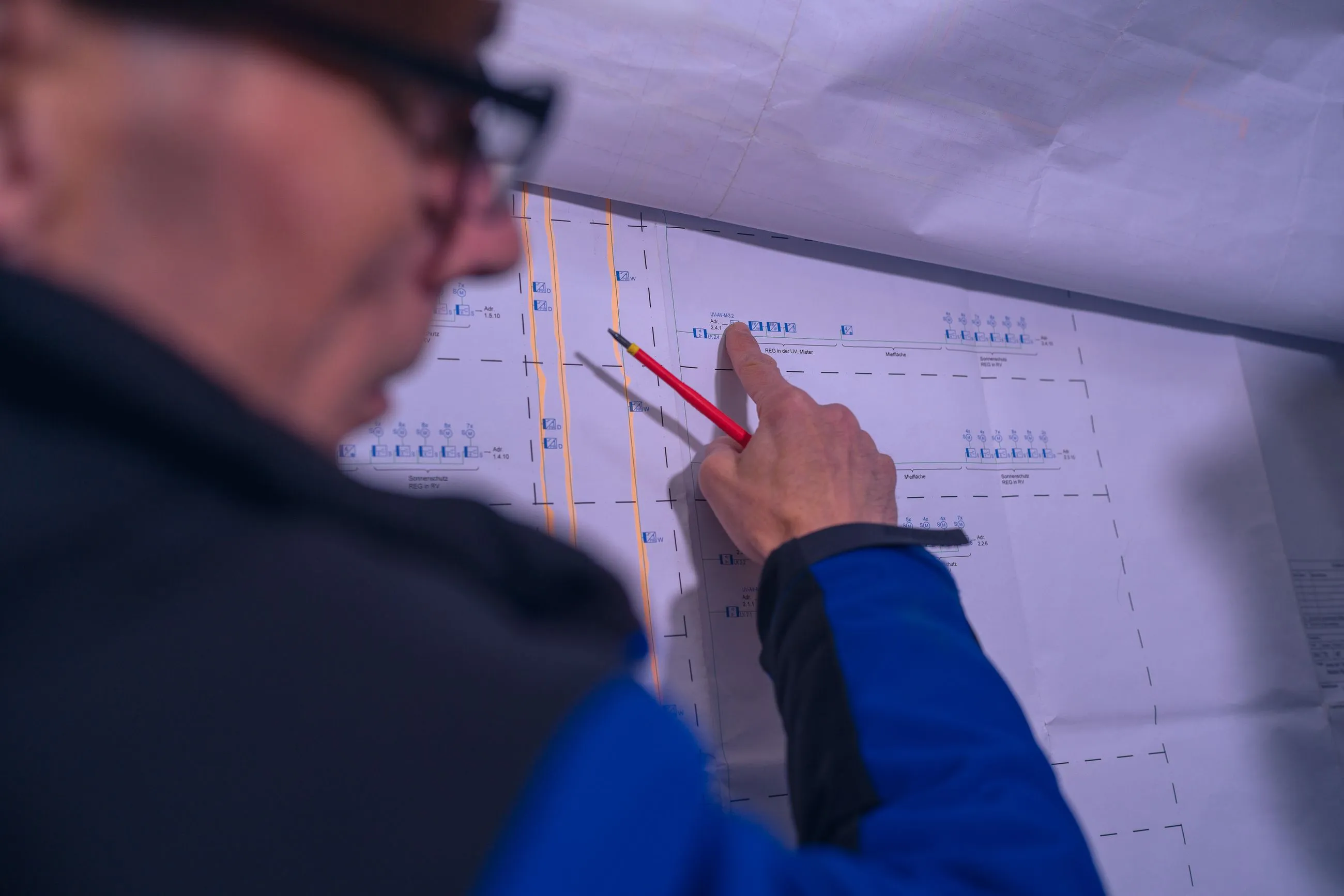 Monteur zeigt mit rotem Stift auf einen technischen Plan oder eine Bauzeichnung an der Wand.
