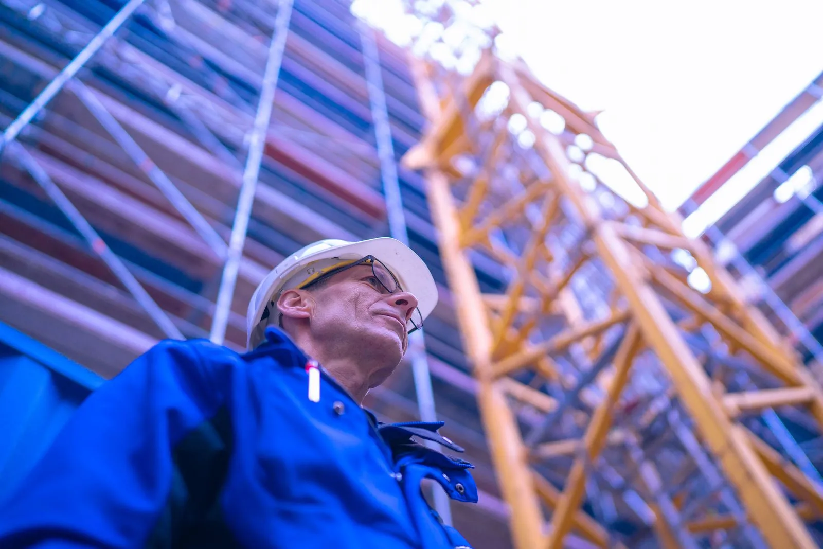 Monteur von IB Walther mit weißem Helm und blauer Arbeitskleidung blickt auf eine Baustelle mit Baukran und Gerüst.