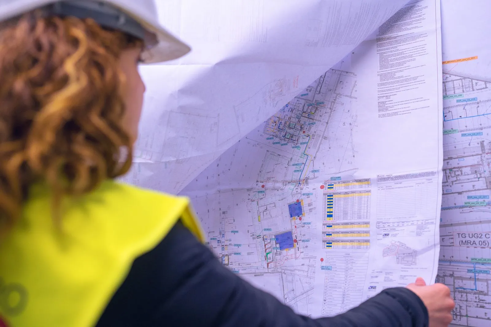 Technikerin mit Helm gelber Weste betrachtet einen detaillierten Bauplan oder technische Zeichnung.