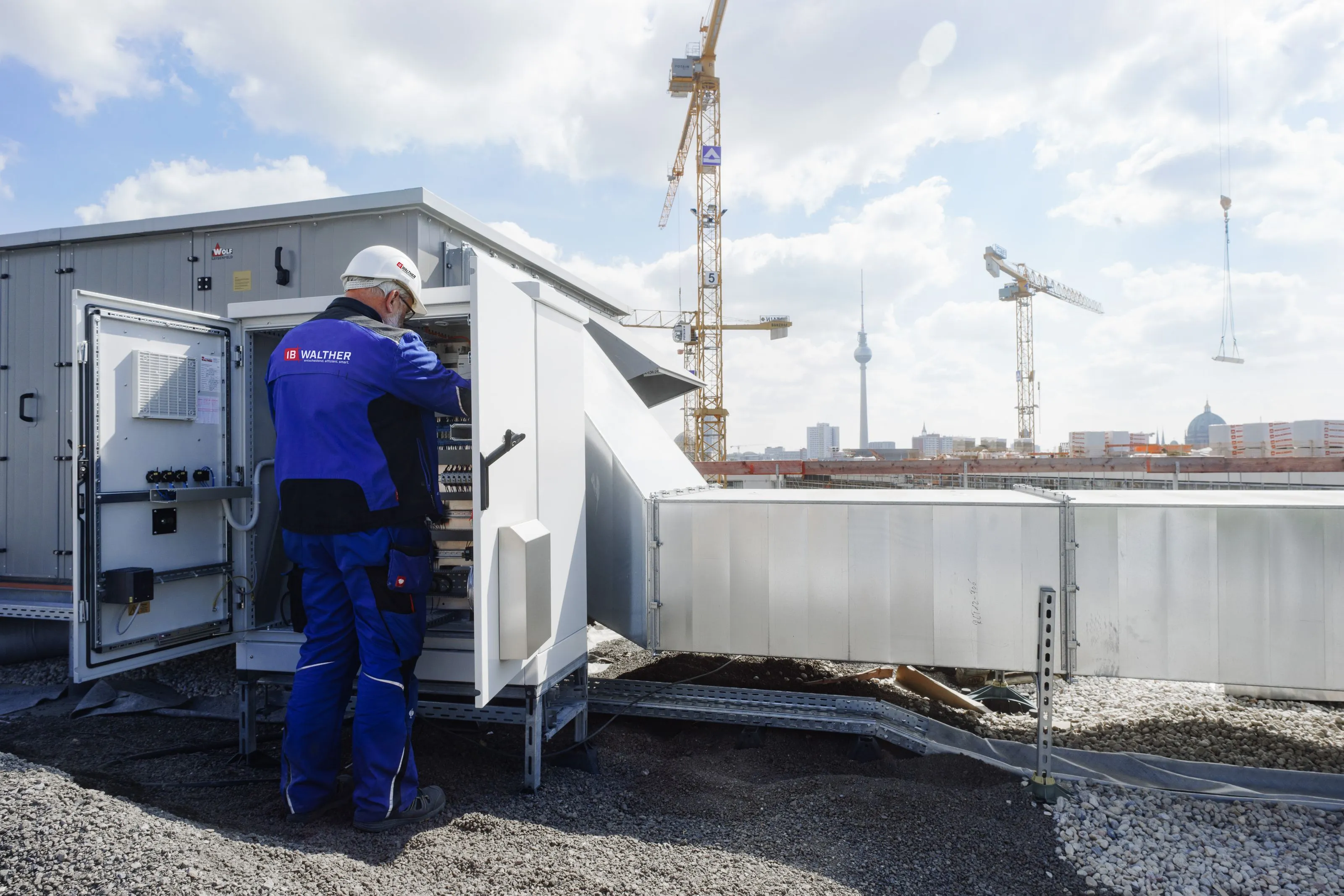 Techniker von IB Walther aus Berlin in blauer Arbeitskleidung mit Helm arbeitet an offenem Schaltschrank auf einer Baustelle mit Kränen im Hintergrund.