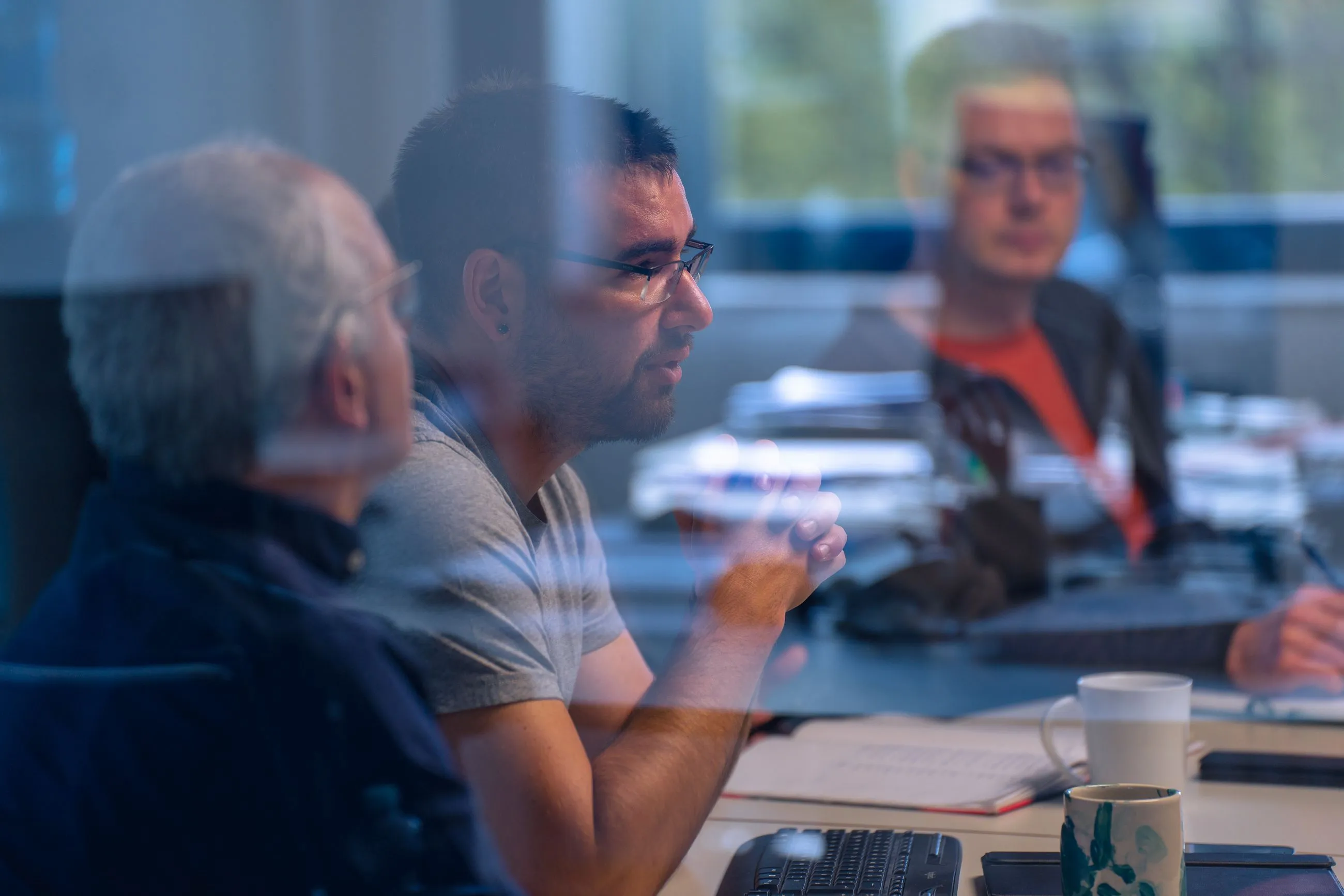 Mitarbeiter im Gespräch bei einem Meeting im Büro, mit Laptop, Notizbuch und Kaffeetassen auf dem Tisch.