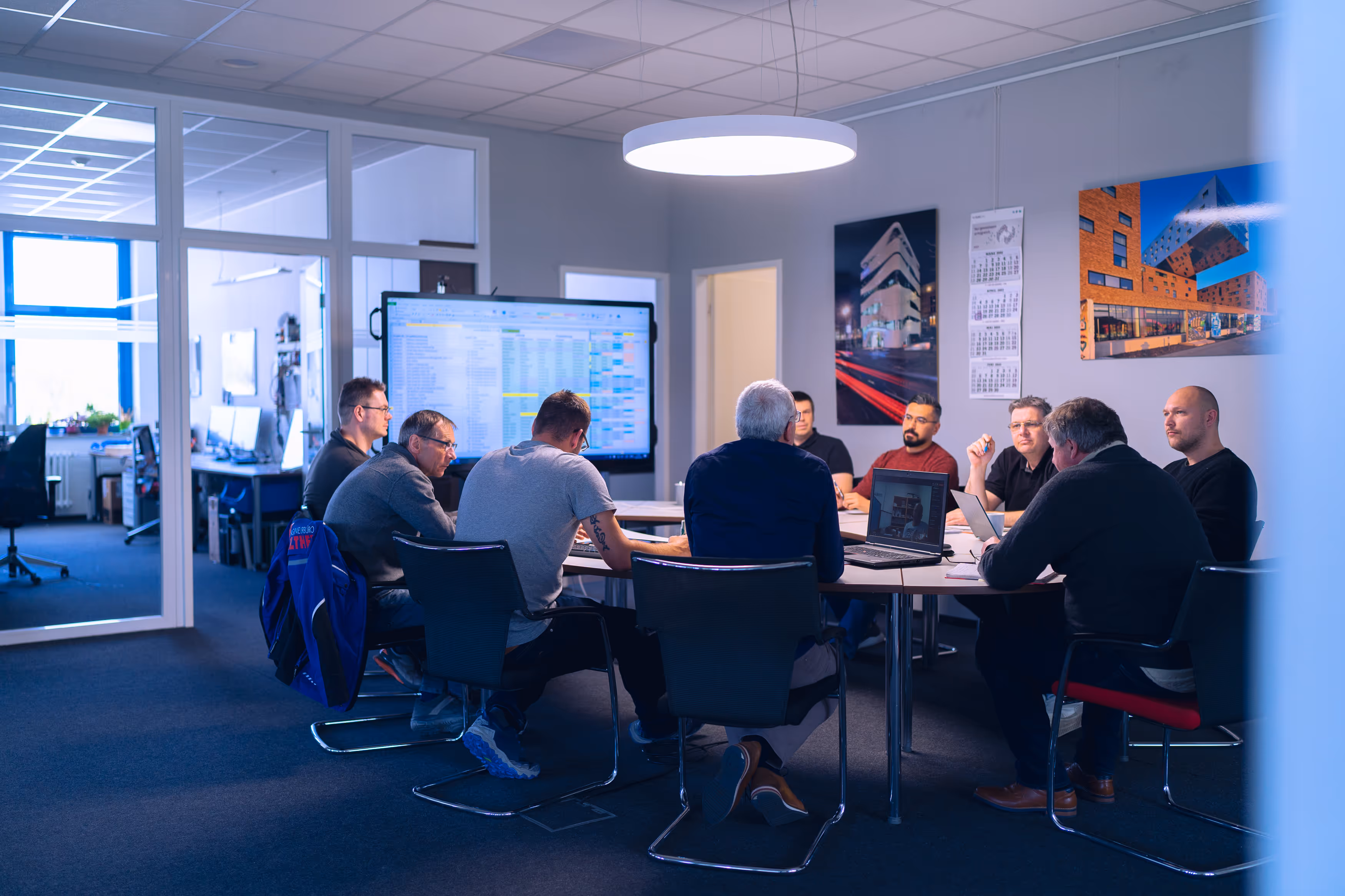 Mitarbeiter von IB Walther die in einem modernen Konferenzraum an einem runden Tisch sitzen und an Laptops und einem großen Bildschirm mit Tabellen arbeiten.