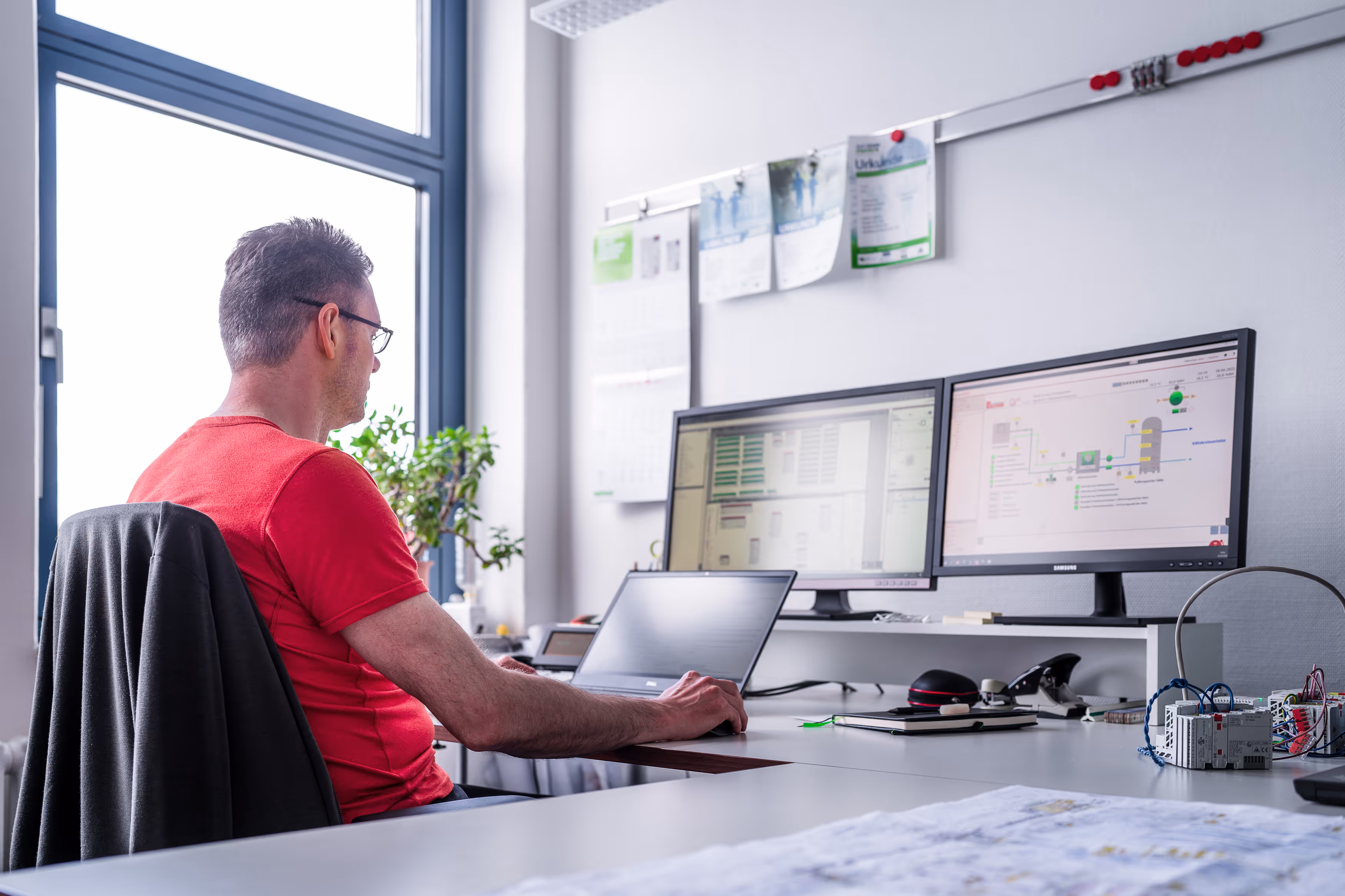IB Walther Mitarbeiter mit rotem T-Shirt arbeitet an einem Laptop mit zwei Monitoren in einem Büro mit technischen Diagrammen.