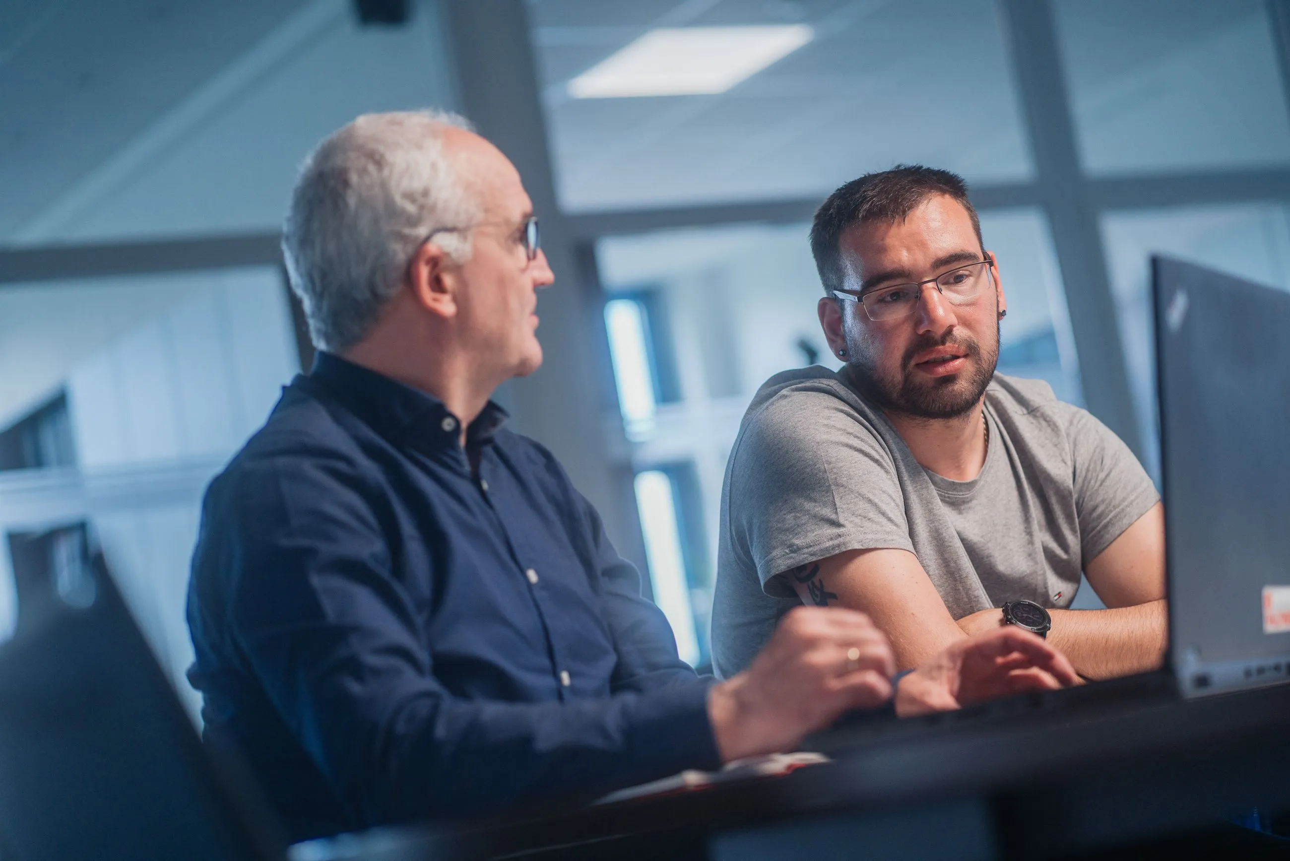 Zwei Mitarbeiter von IB Walther sitzen an einem Tisch und schauen auf einen Laptop-Bildschirm, einer trägt ein graues T-Shirt, der andere ein dunkles Hemd.